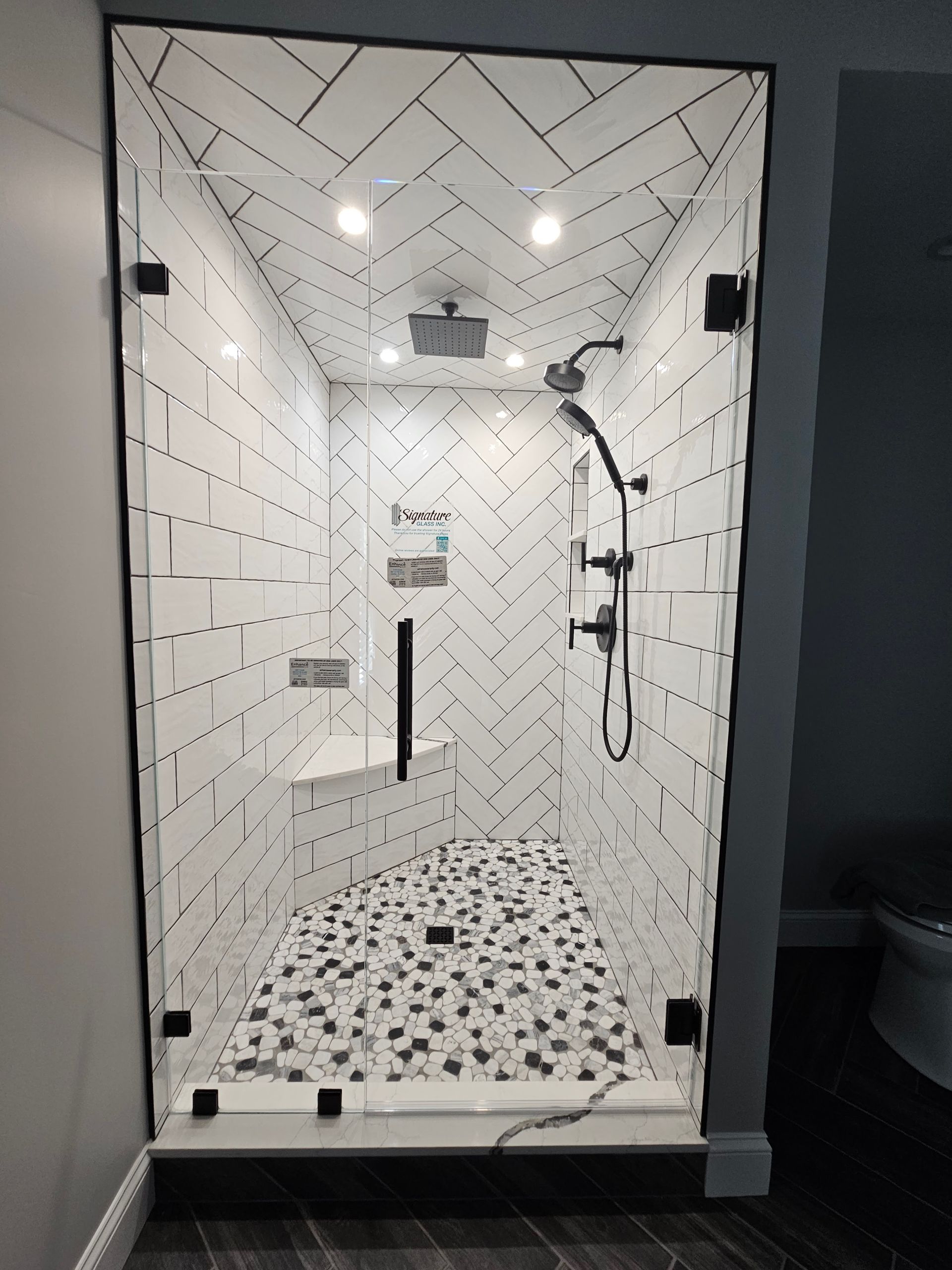 Glass-enclosed shower with white herringbone tile walls, black fixtures, and mosaic floor.
