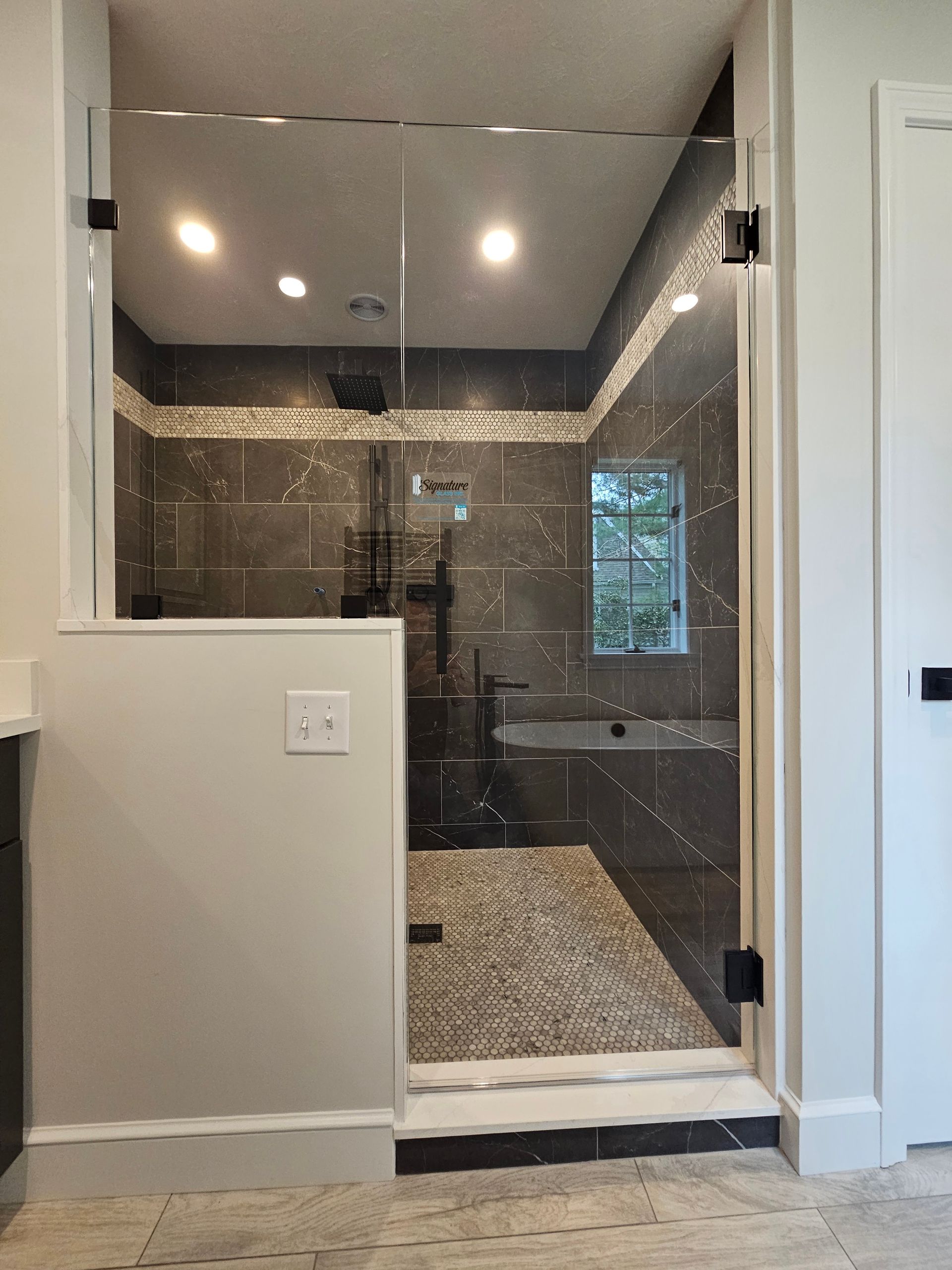 Shower with glass door, black fixtures, gray and tan tiled walls, and pebble floor.