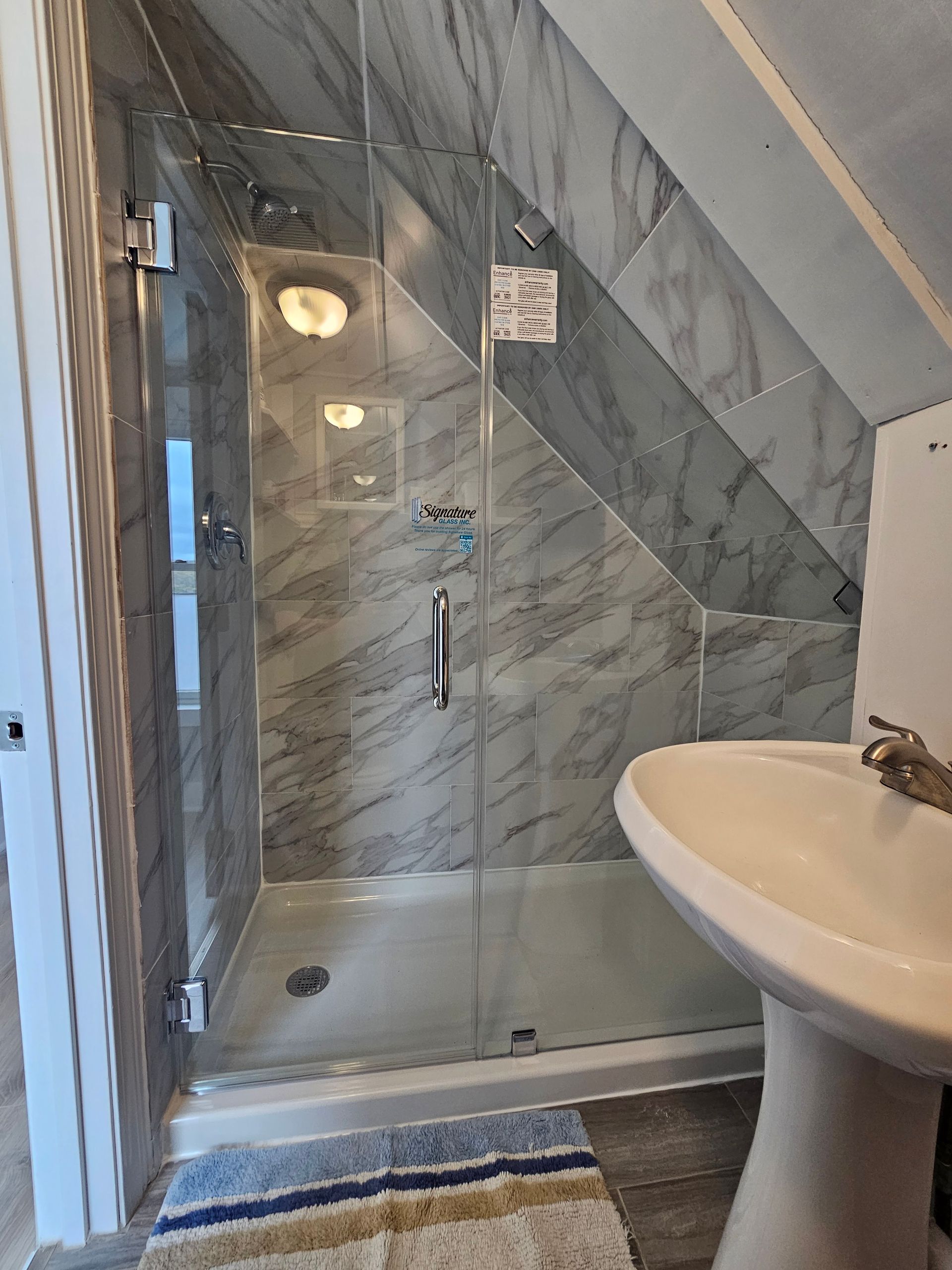 Shower stall with marble-patterned walls and glass door, next to a pedestal sink in a bathroom.