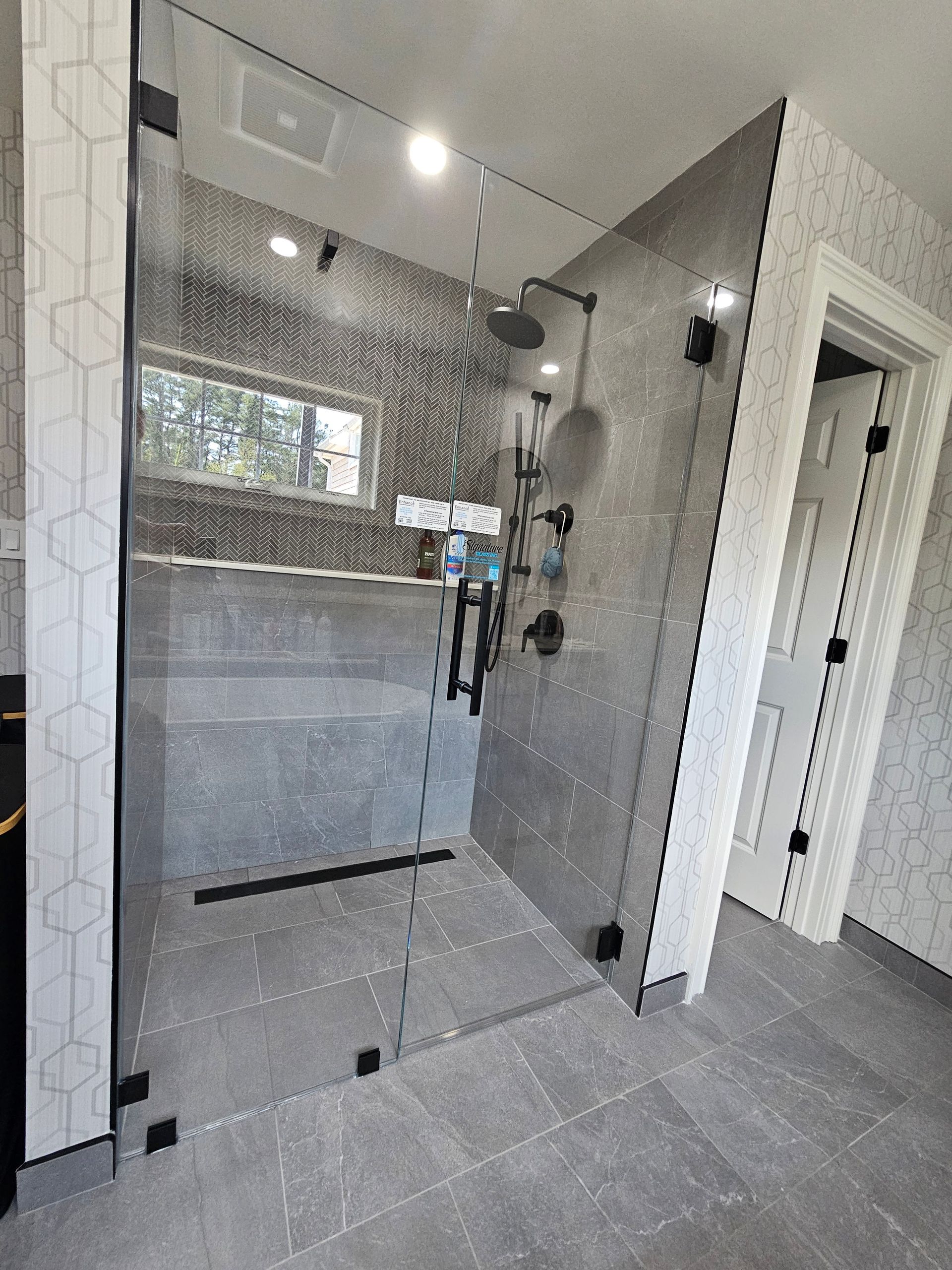 Modern bathroom with a glass shower enclosure, gray tile, black fixtures, and a window.