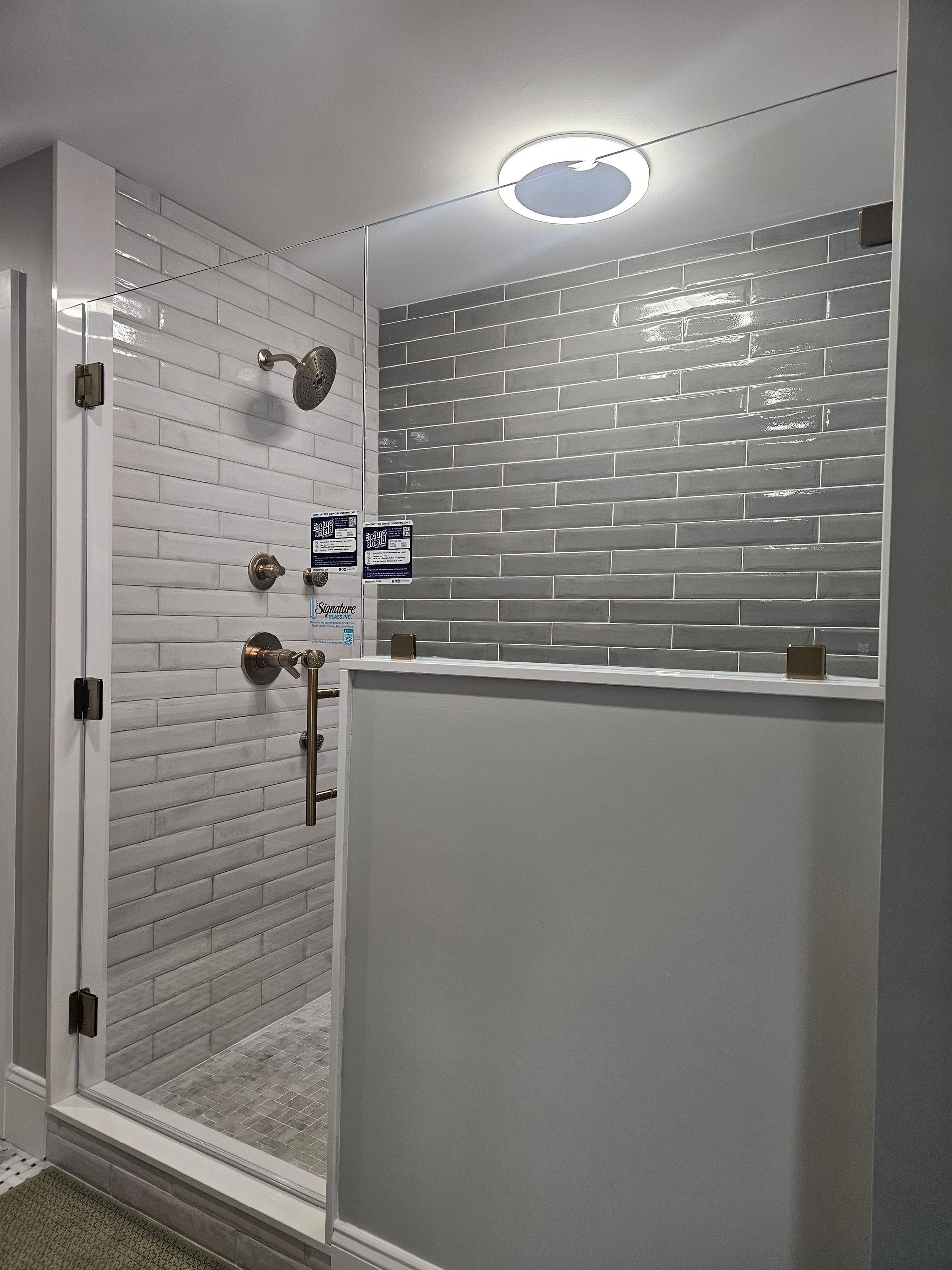 Modern shower with glass doors, white brick-like tiles, bronze fixtures, and a circular ceiling light.