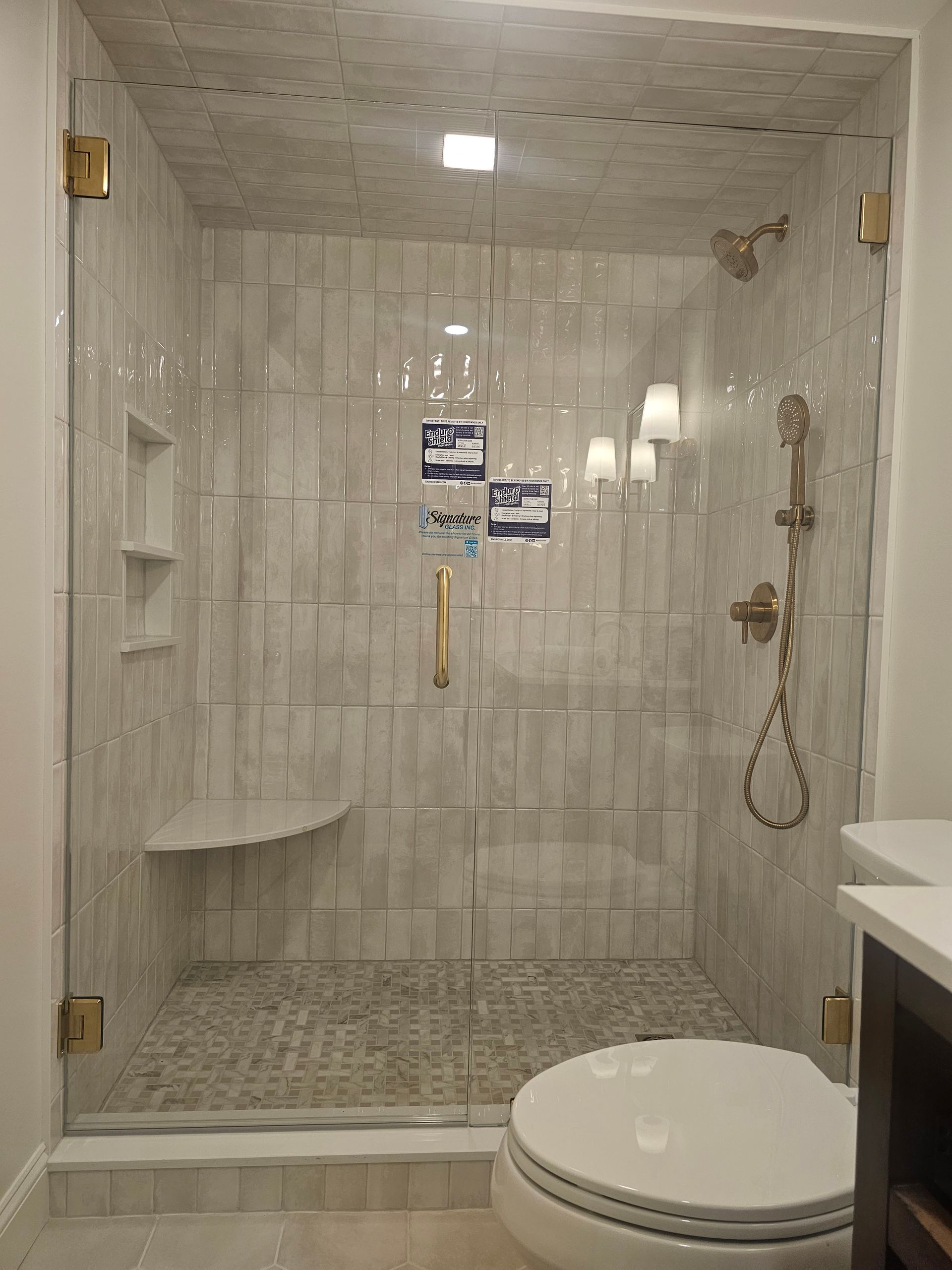 Glass-door shower with gold fixtures, tiled walls, corner seat, and built-in shelf. Next to a toilet in a bathroom.