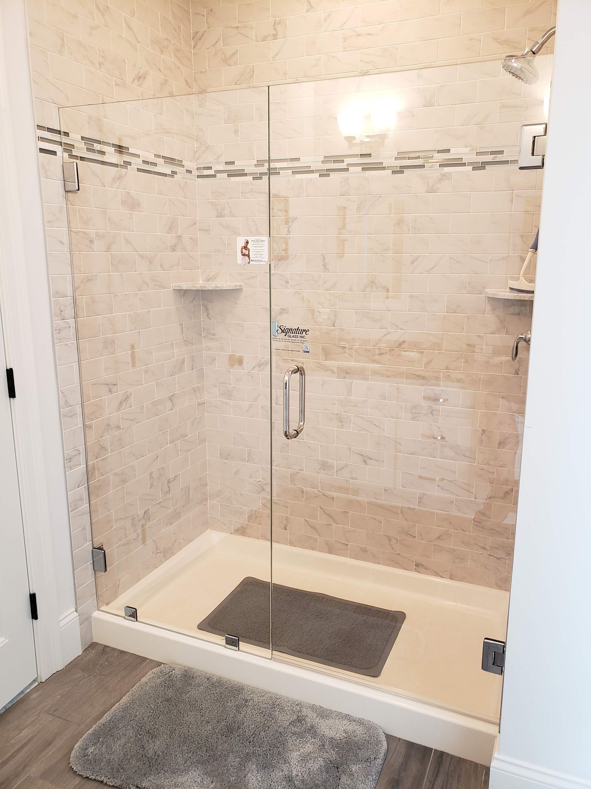 Shower stall with glass door, beige tiled walls, white base, and gray rug on a light wood floor.