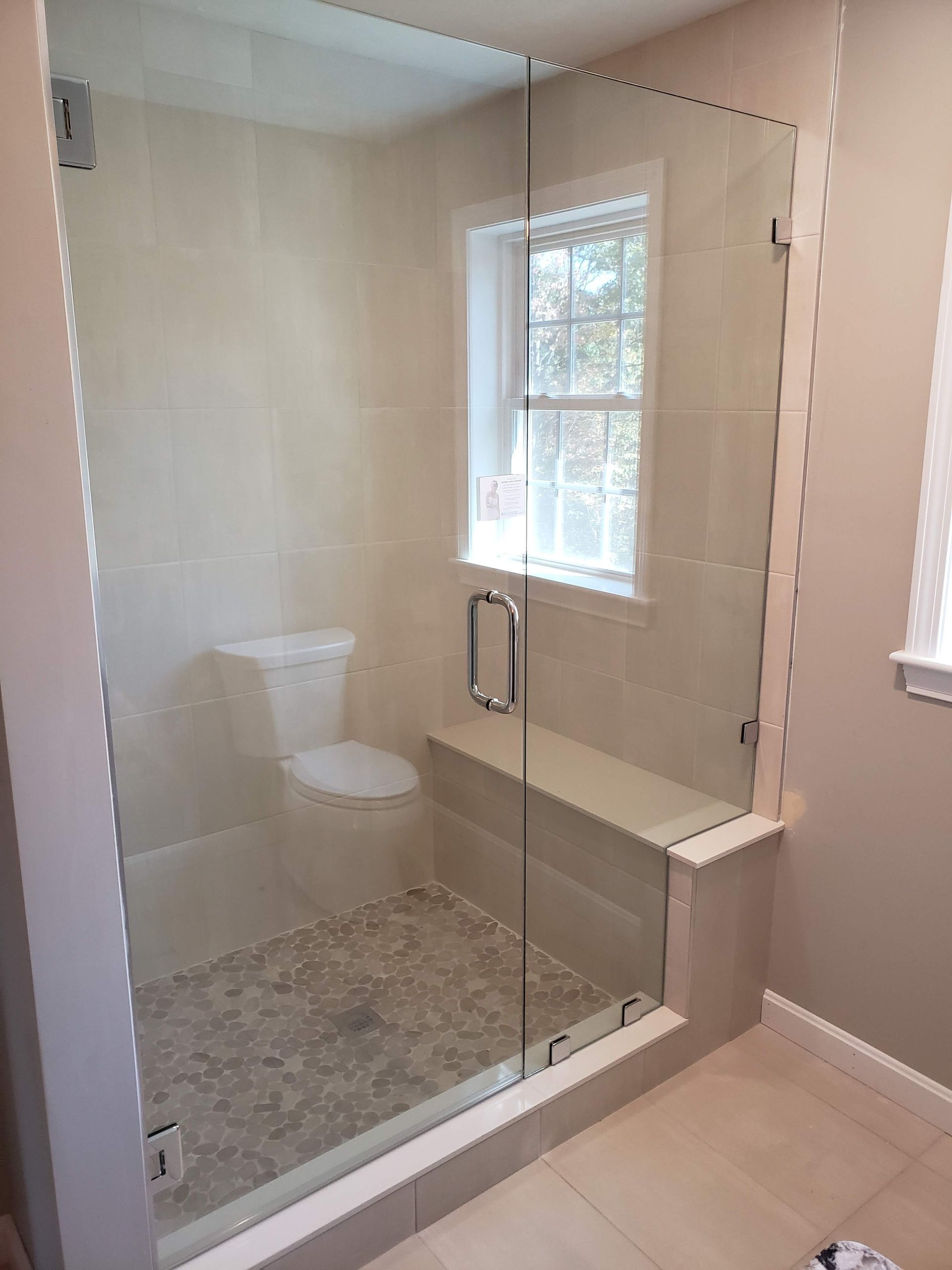Glass shower enclosure with a bench, toilet, and window, tiled walls and pebble floor.