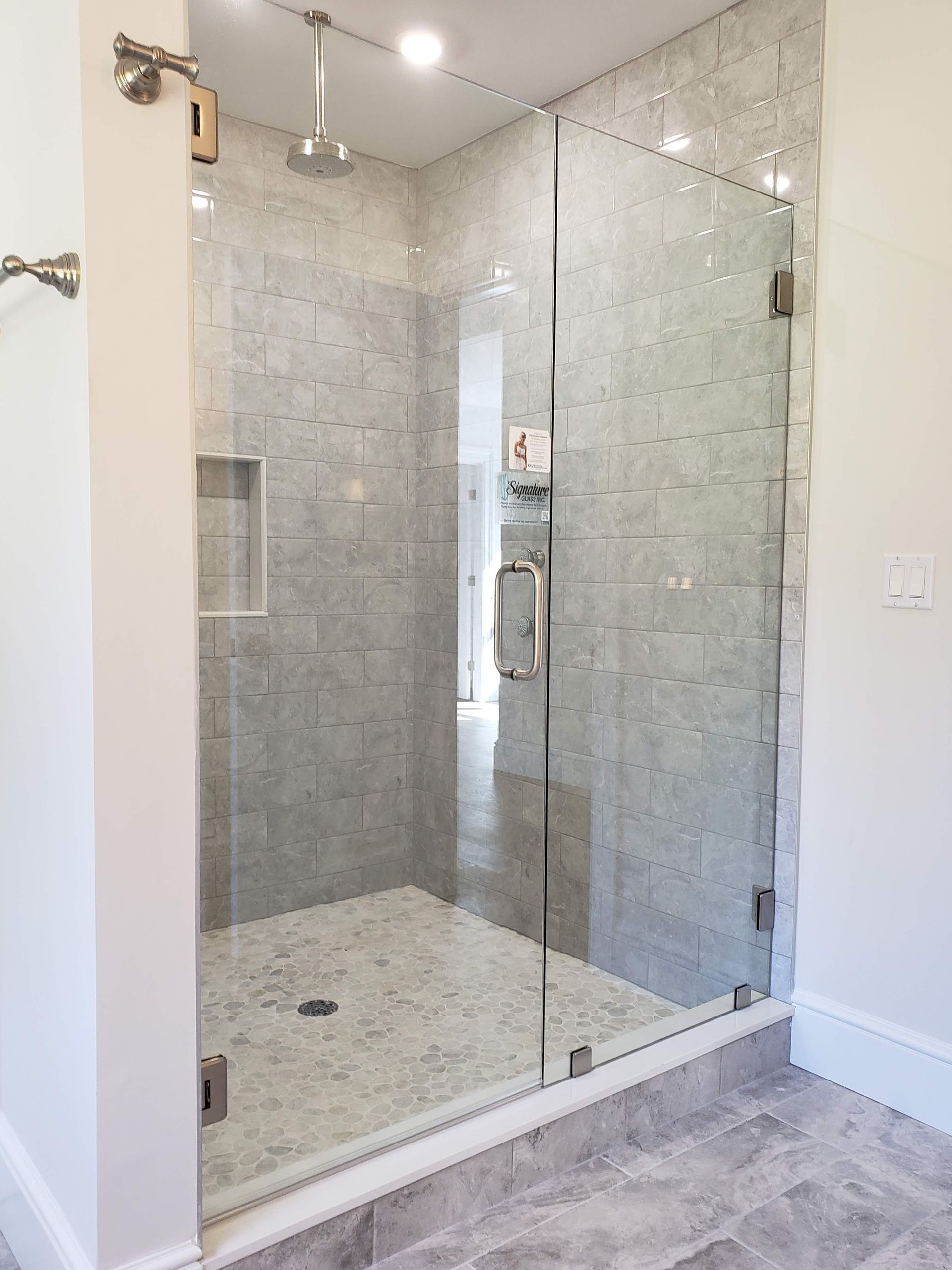Glass-enclosed shower with gray tiled walls and floor, a rainfall shower head, and a stainless steel handle.