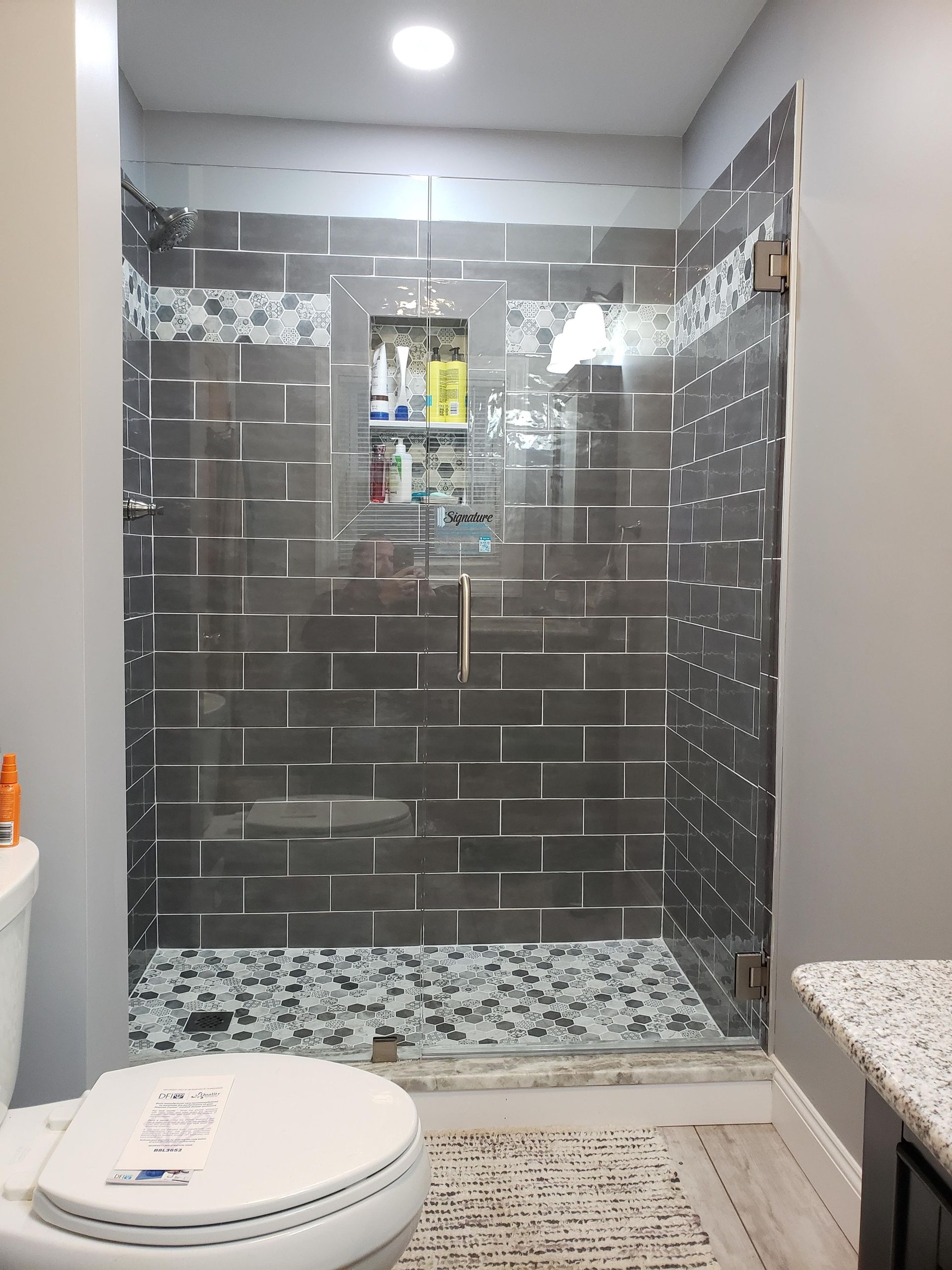 Gray-tiled shower with glass doors, built-in shelf, and small patterned floor, next to a toilet.