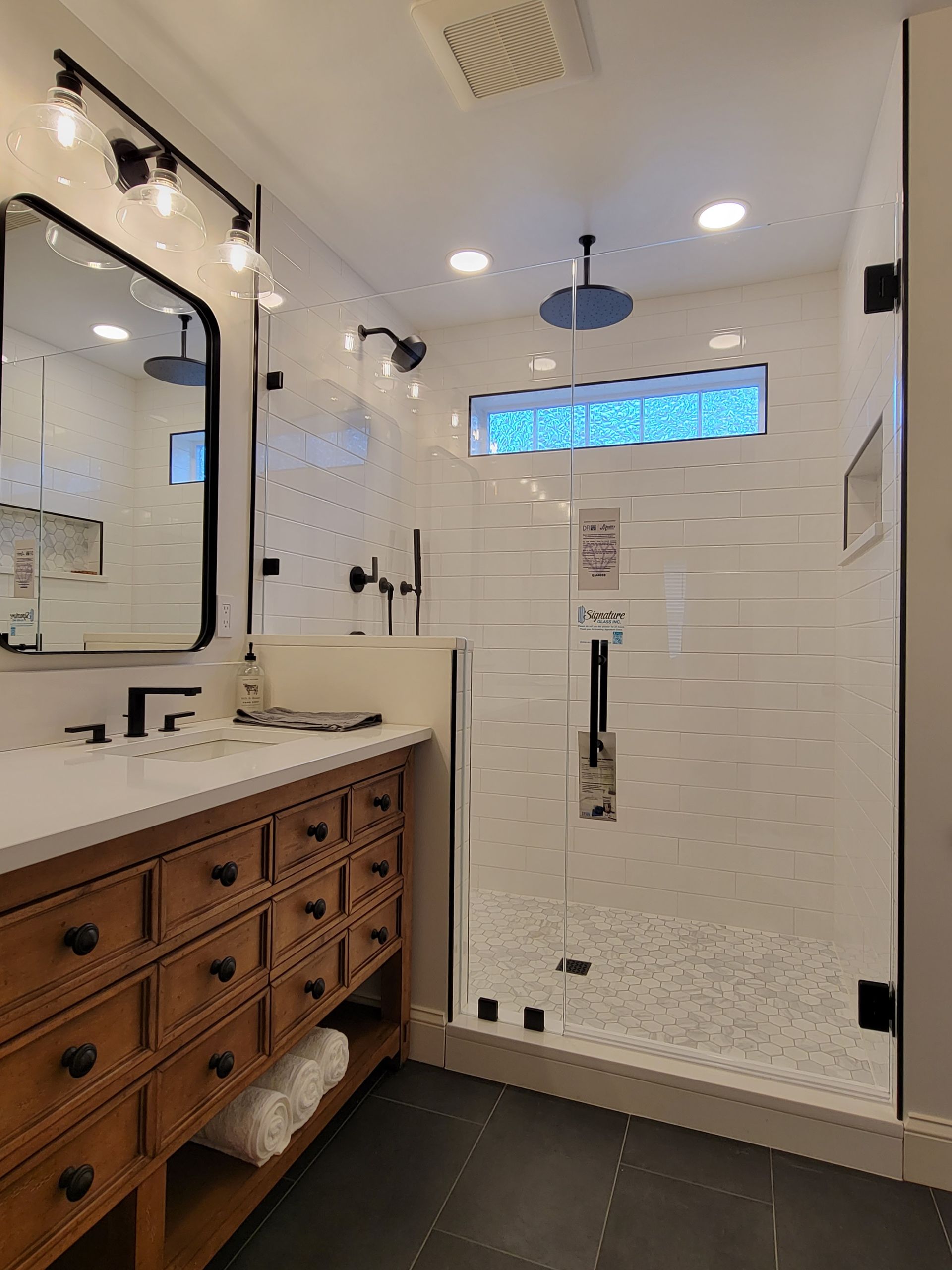 Bathroom with a wooden vanity, a glass shower with a rain showerhead, and white tiled walls.