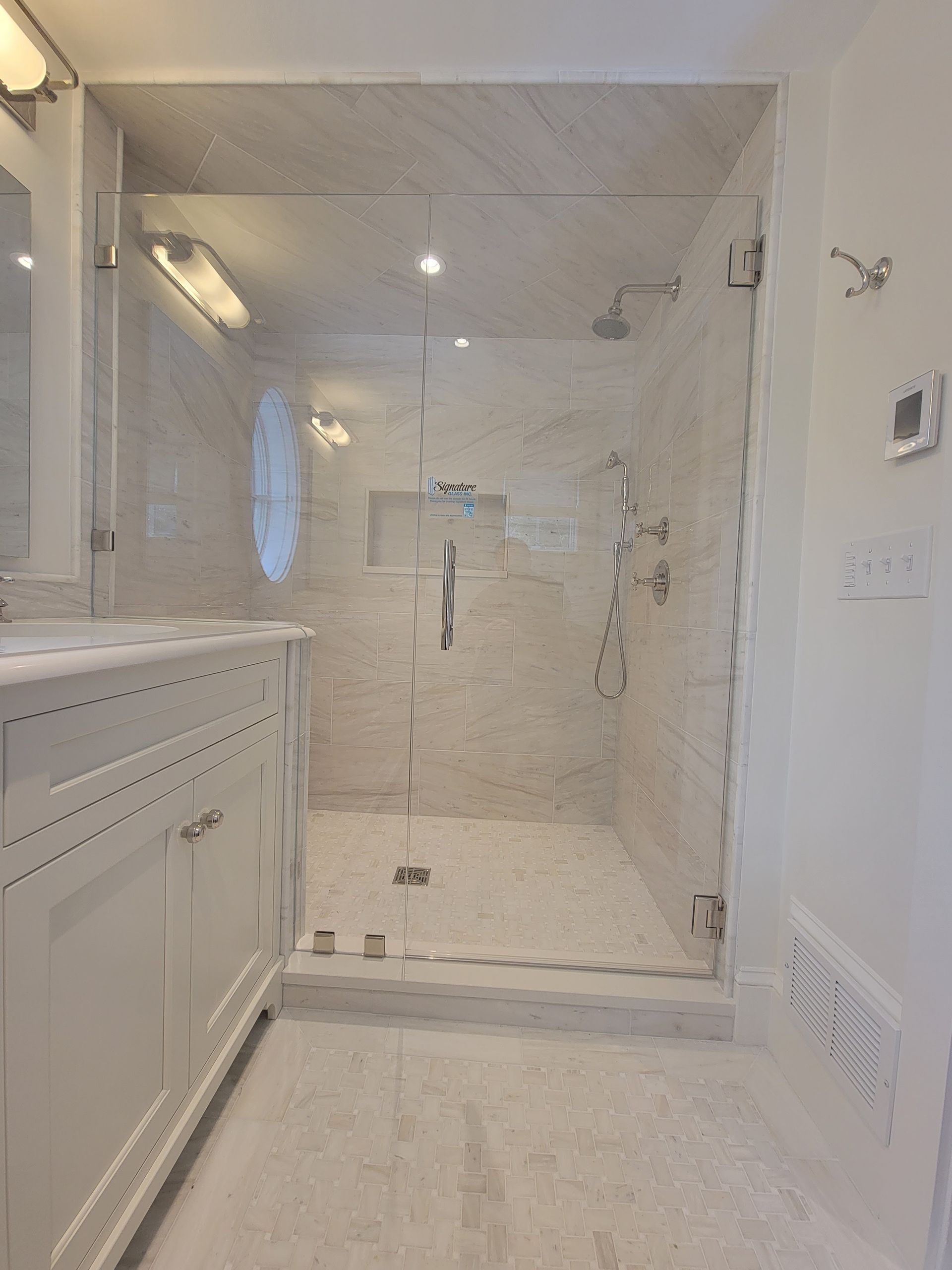 Glass shower enclosure in a white bathroom with marble-look tile and a white vanity.