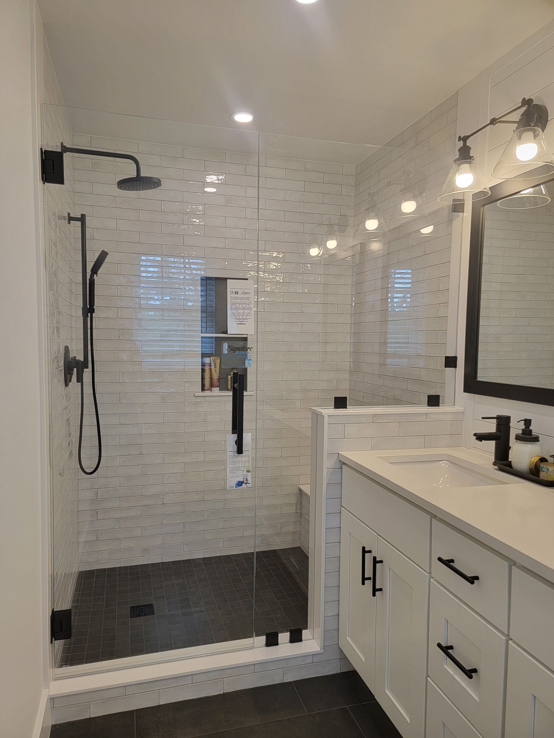 Bathroom with glass shower, white tile walls, dark floor and fixtures, white vanity.