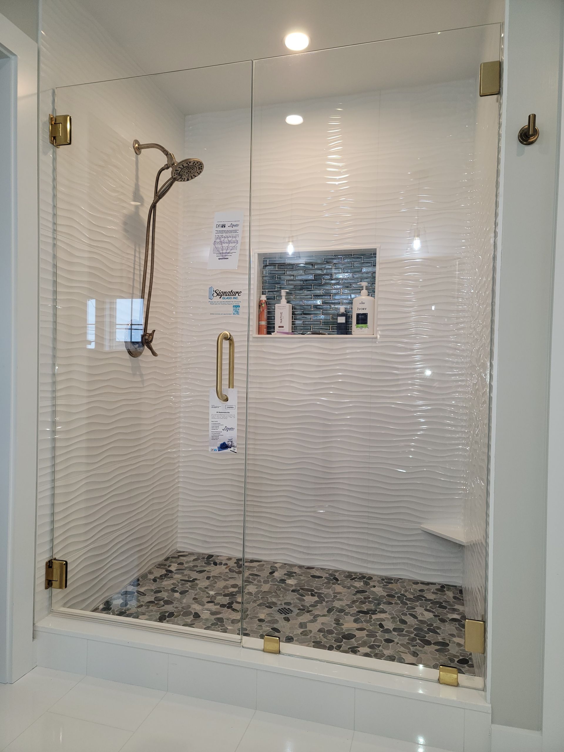 Glass shower enclosure with gold hardware, pebbled floor, and white tiled walls.