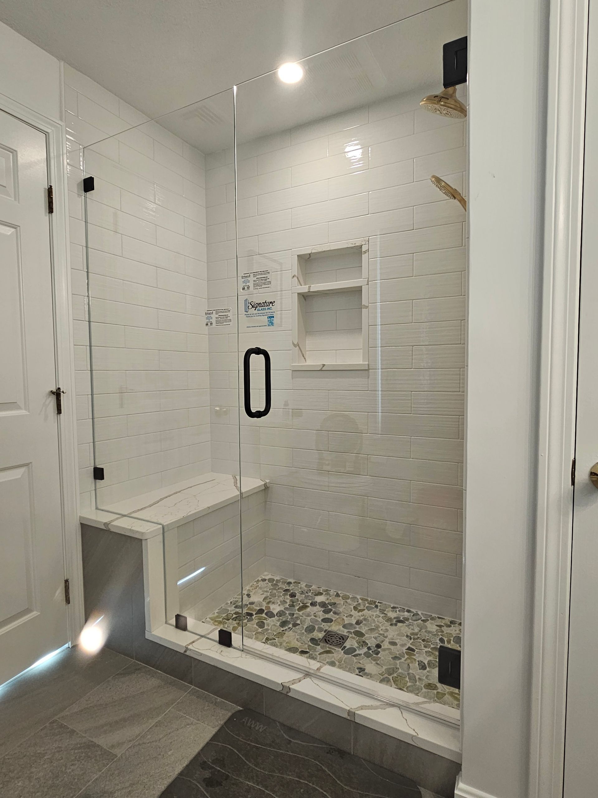 Glass-enclosed shower with white tiled walls, a built-in bench, and pebble floor. Black door handle and fixtures.