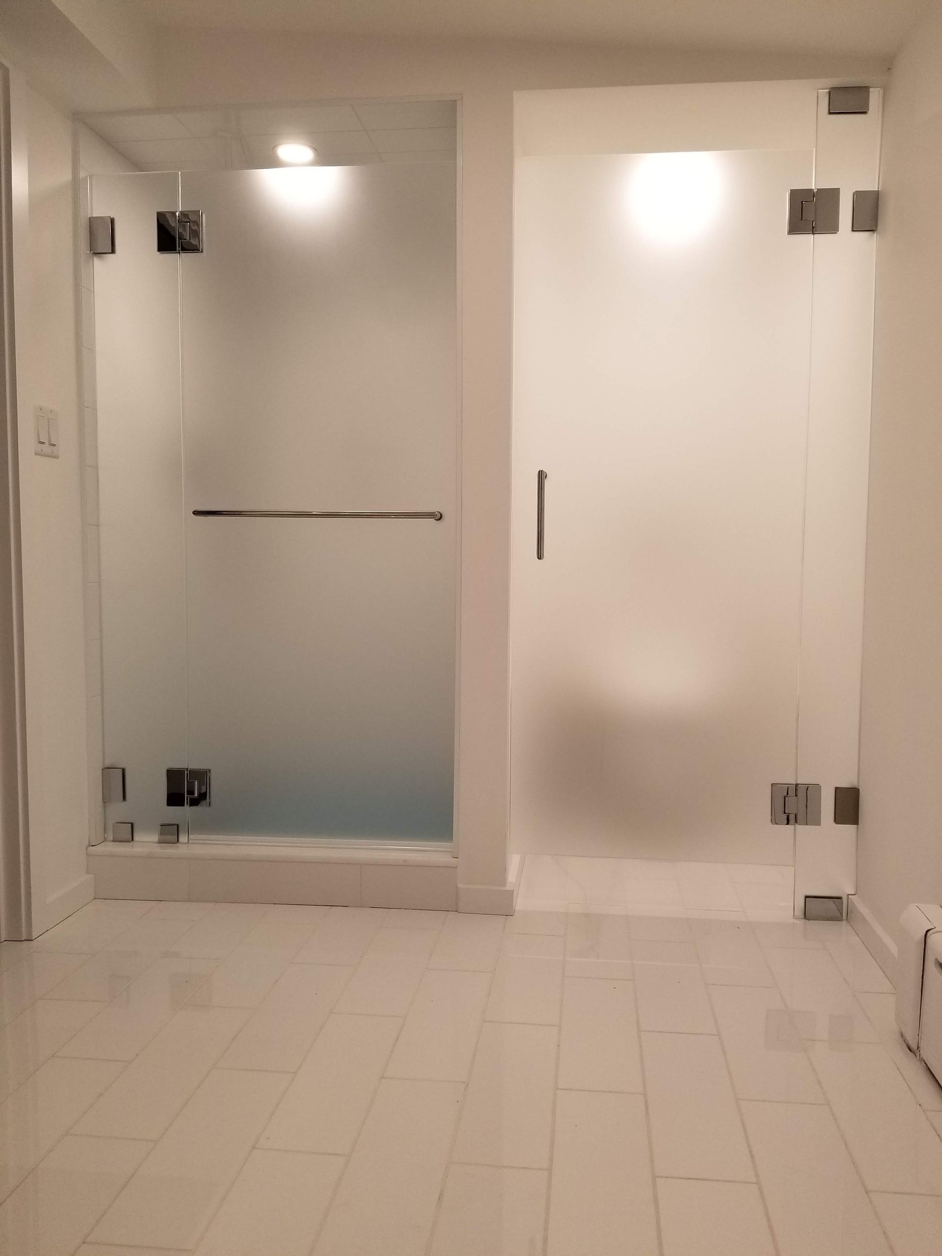 Two frosted glass shower enclosures in a white-tiled bathroom. One has a bar handle, the other a vertical handle.