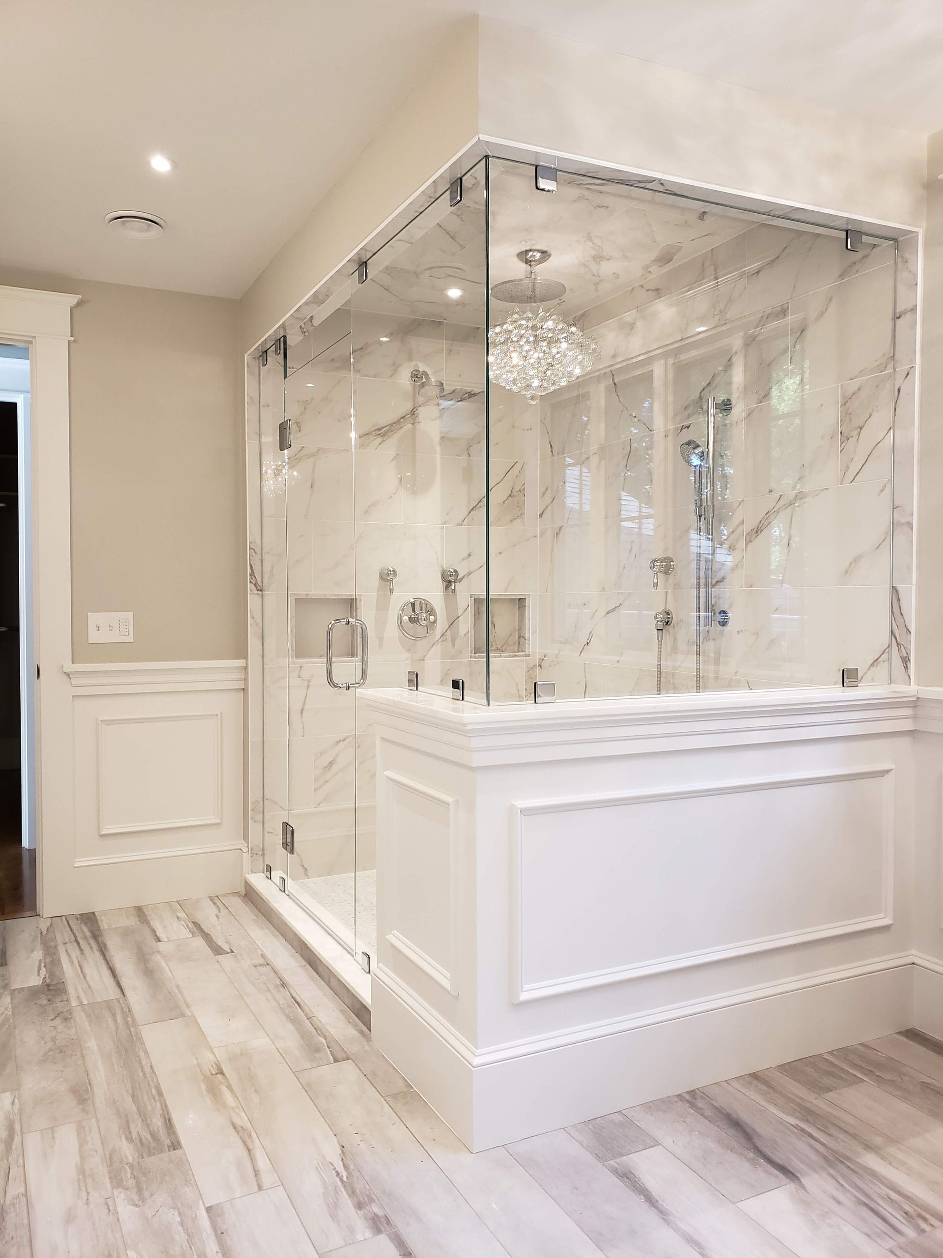Elegant shower with glass walls, marble tile, and white paneling.  Chandelier hangs inside.