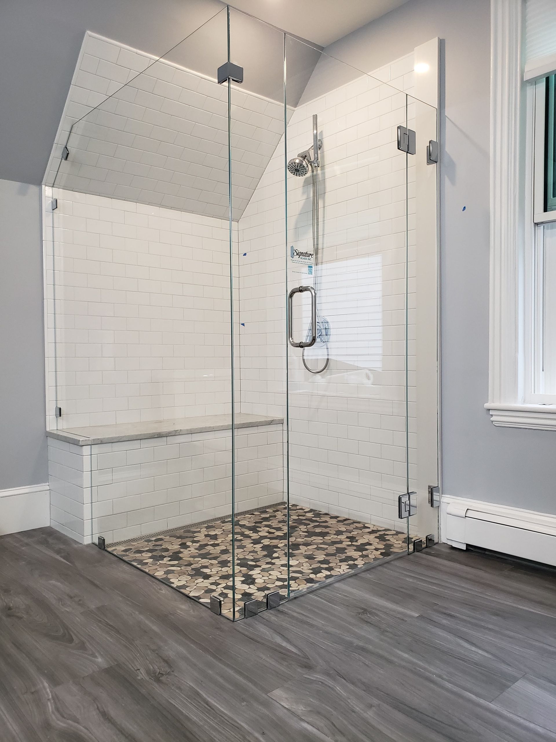 Glass shower with bench and pebble floor, white tile walls, in a bathroom with gray wood-look flooring.