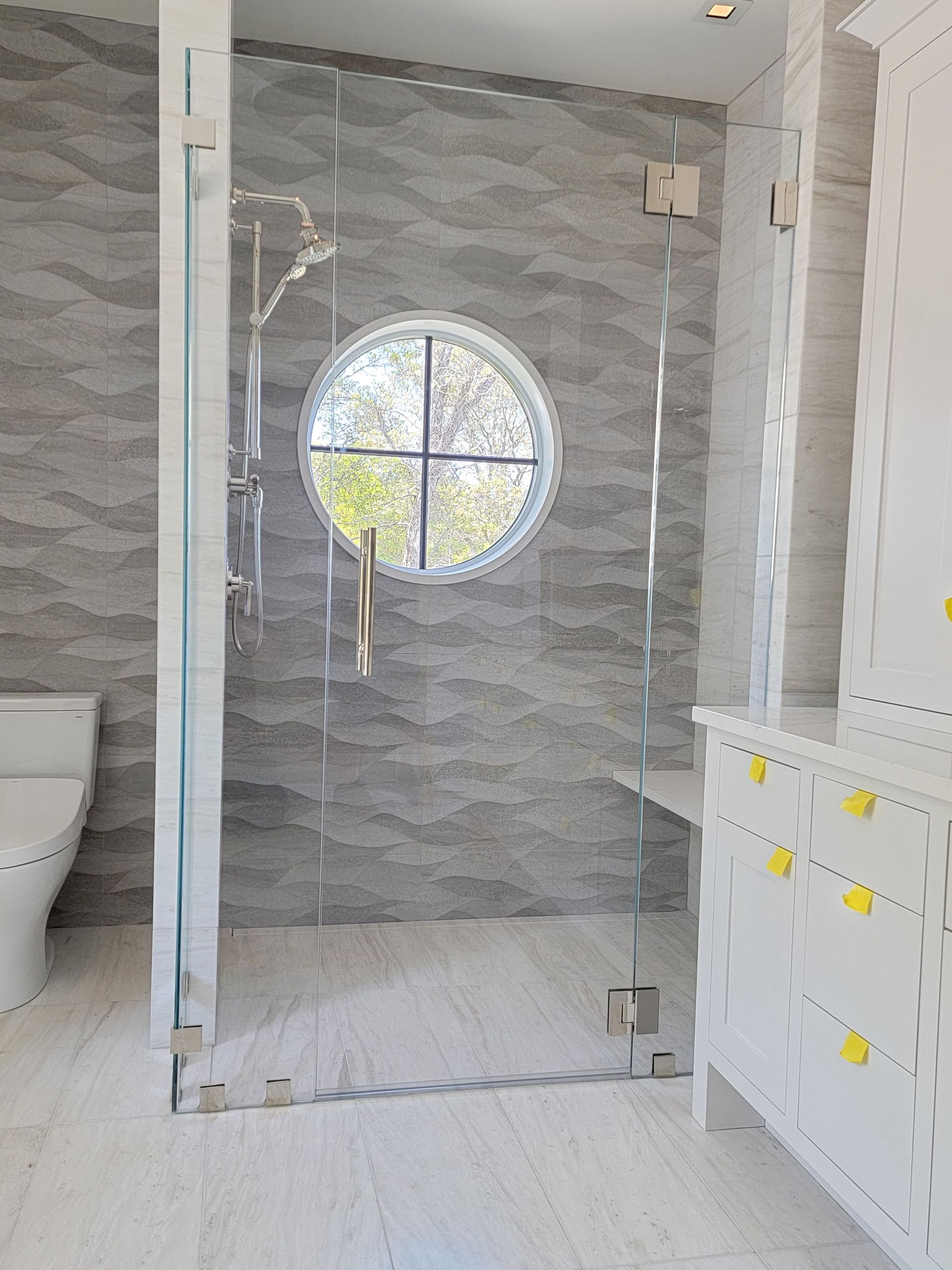Bathroom with gray wave tile, clear glass shower door, and round window. White toilet and cabinet are visible.