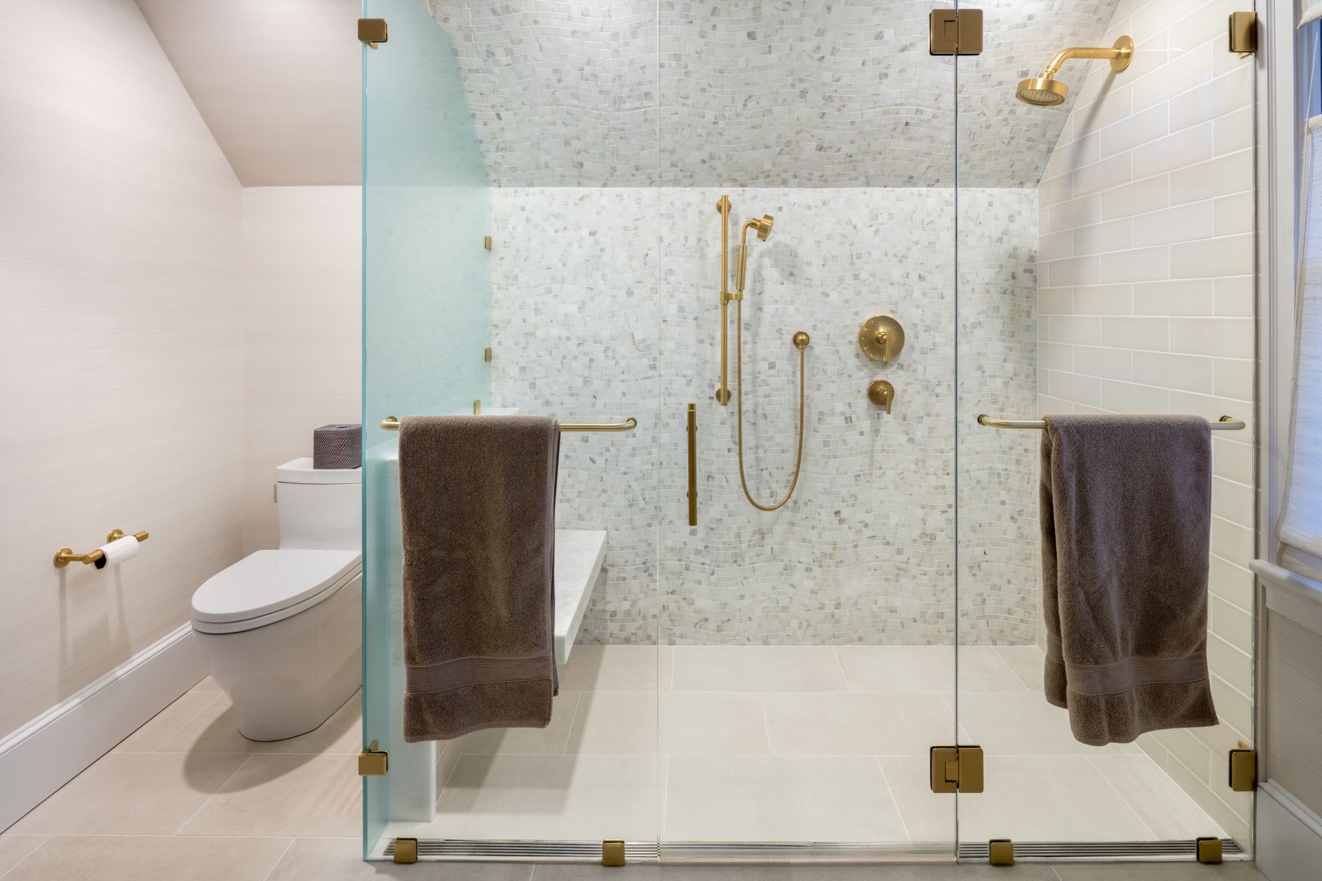 Modern bathroom with glass shower enclosure, gold fixtures, and a white toilet.  Two brown towels hang on the shower.