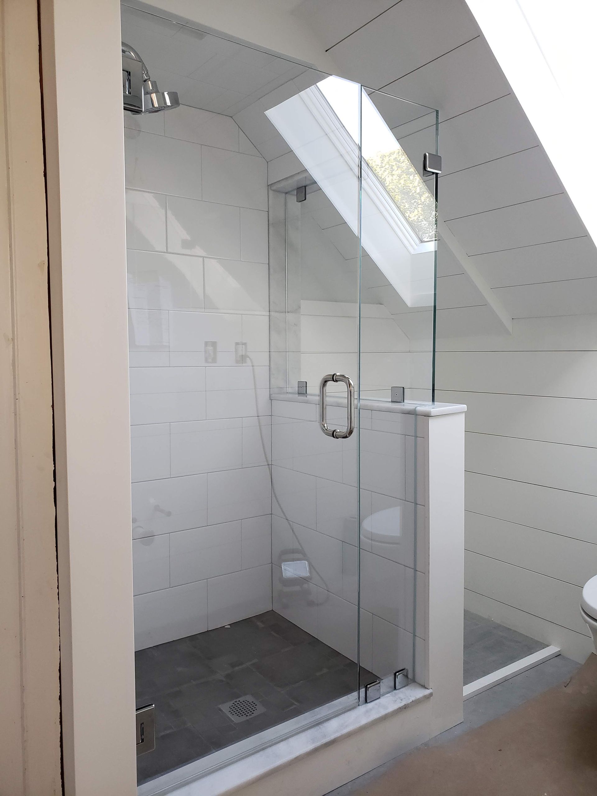 Shower with glass door, white tiled walls, and skylight. Gray floor, chrome fixtures.