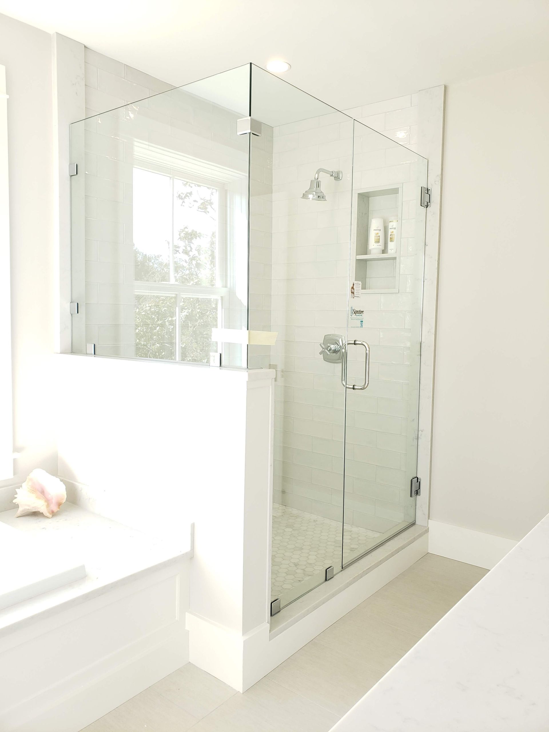 Glass-walled shower with white tile interior, next to a window and white walls.