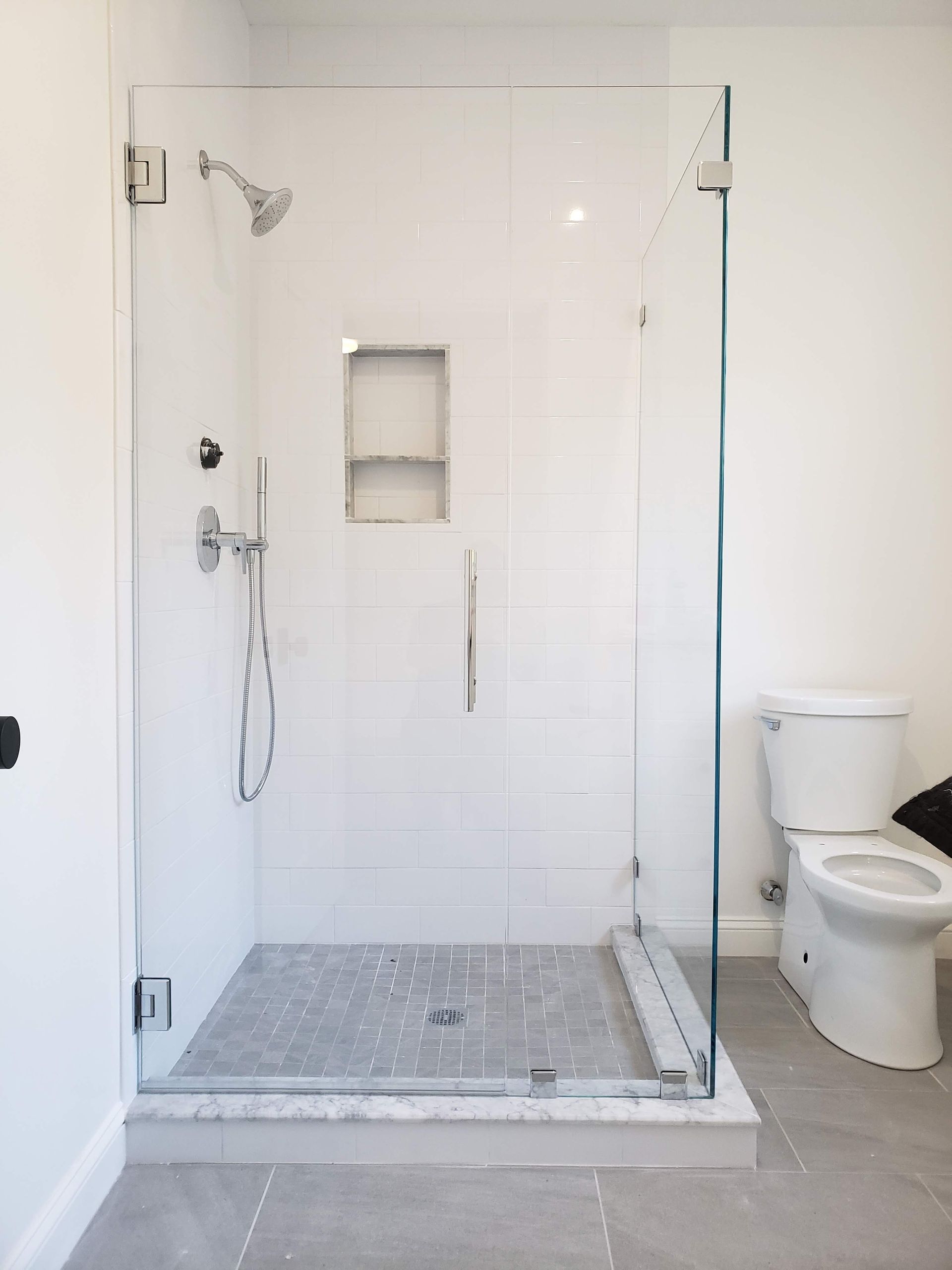 Glass shower with white tile walls and gray stone floor, next to a toilet.