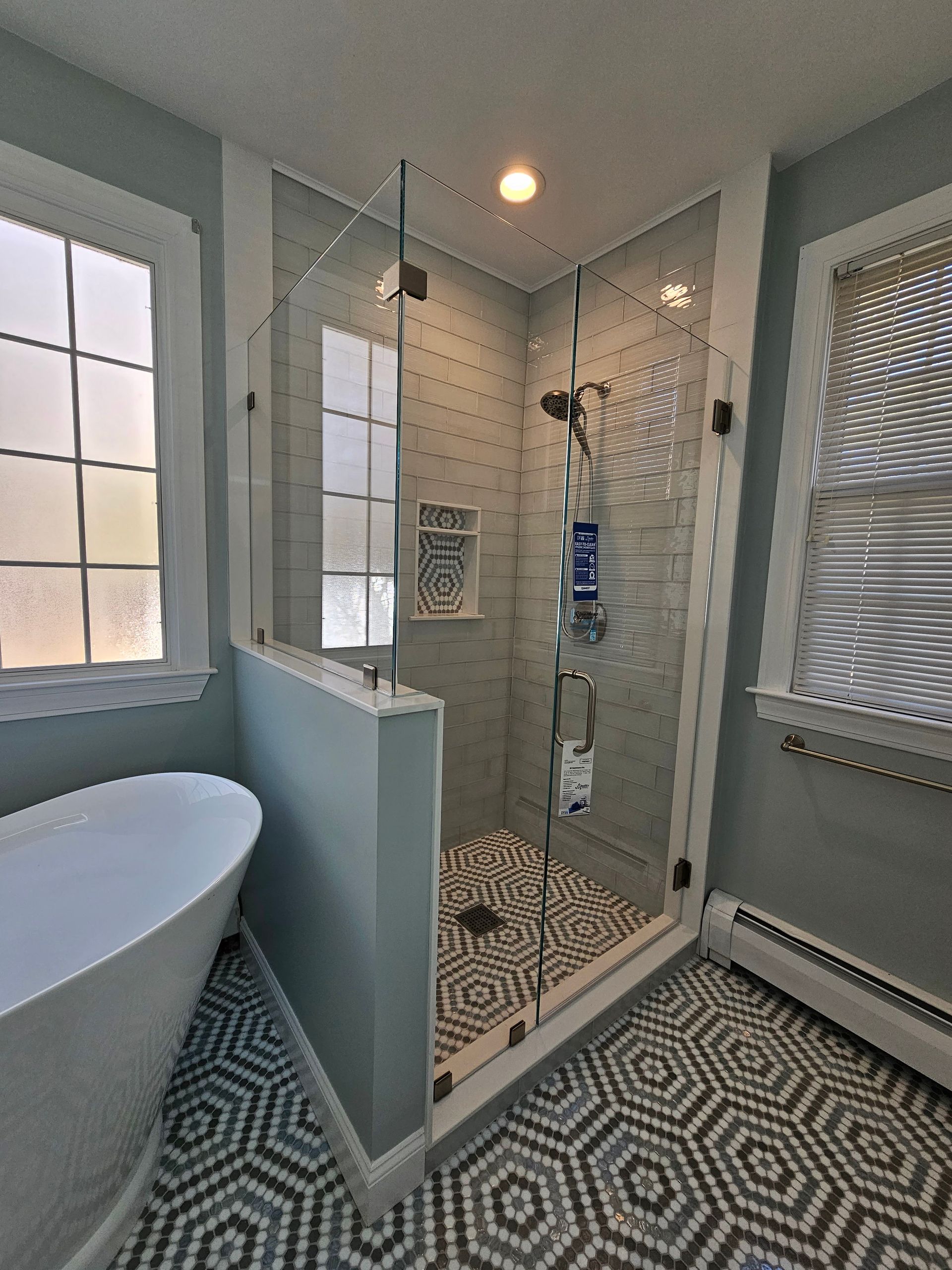 Bathroom with glass shower, white tub, and patterned floor. Light blue walls and natural light.