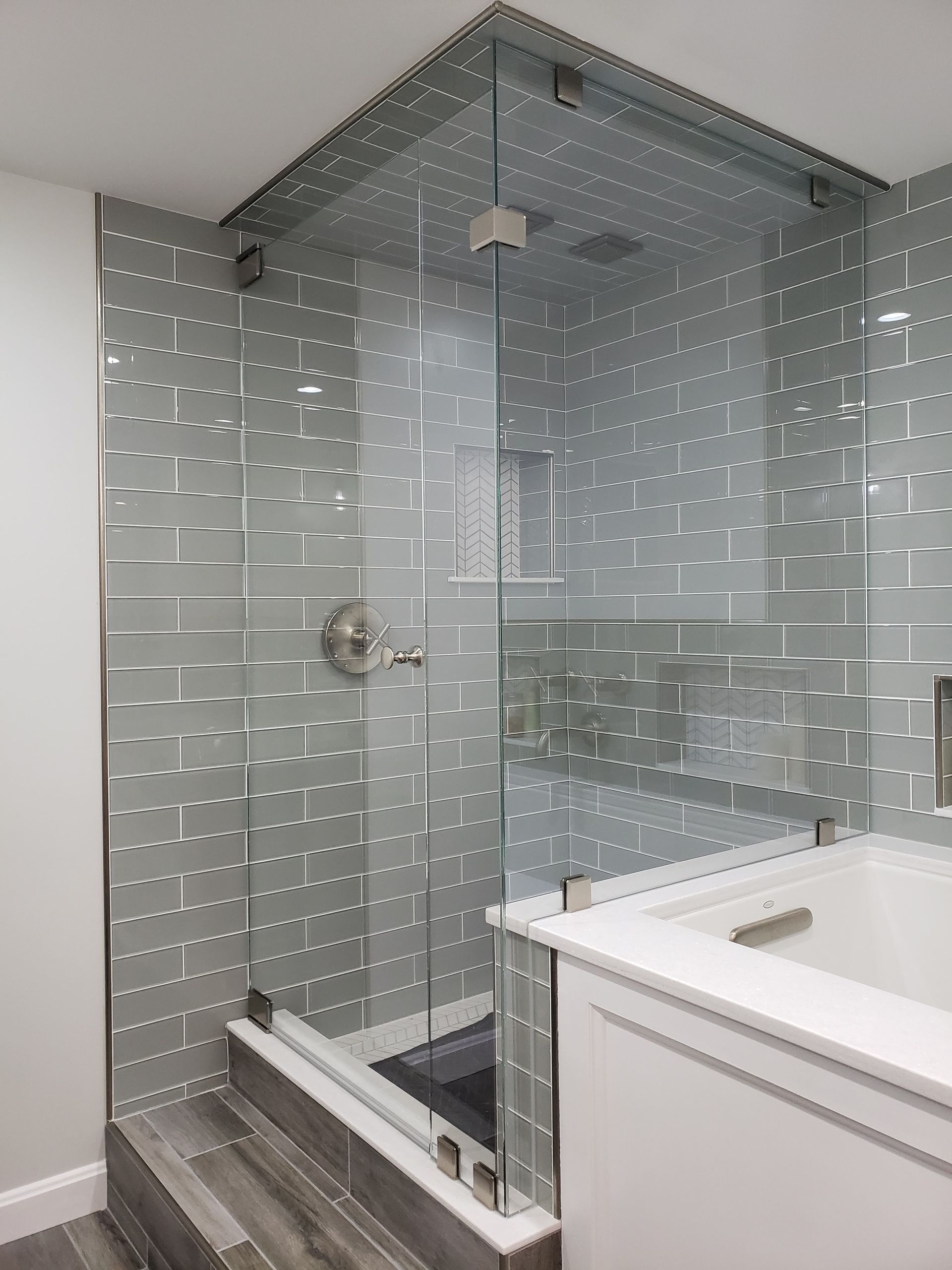 Corner shower with glass doors, gray tiled walls, silver fixtures, and a white vanity next to a bathtub.