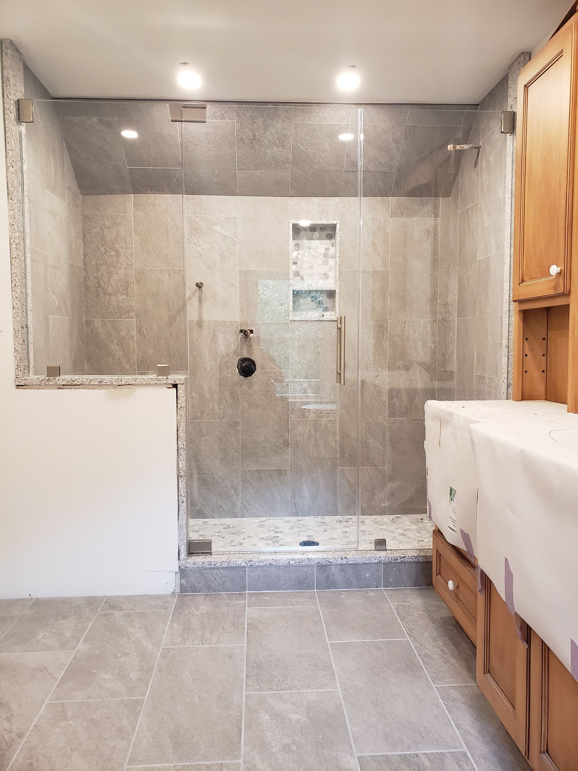 New shower stall with gray tiled walls and glass doors; unfinished cabinet on the right.