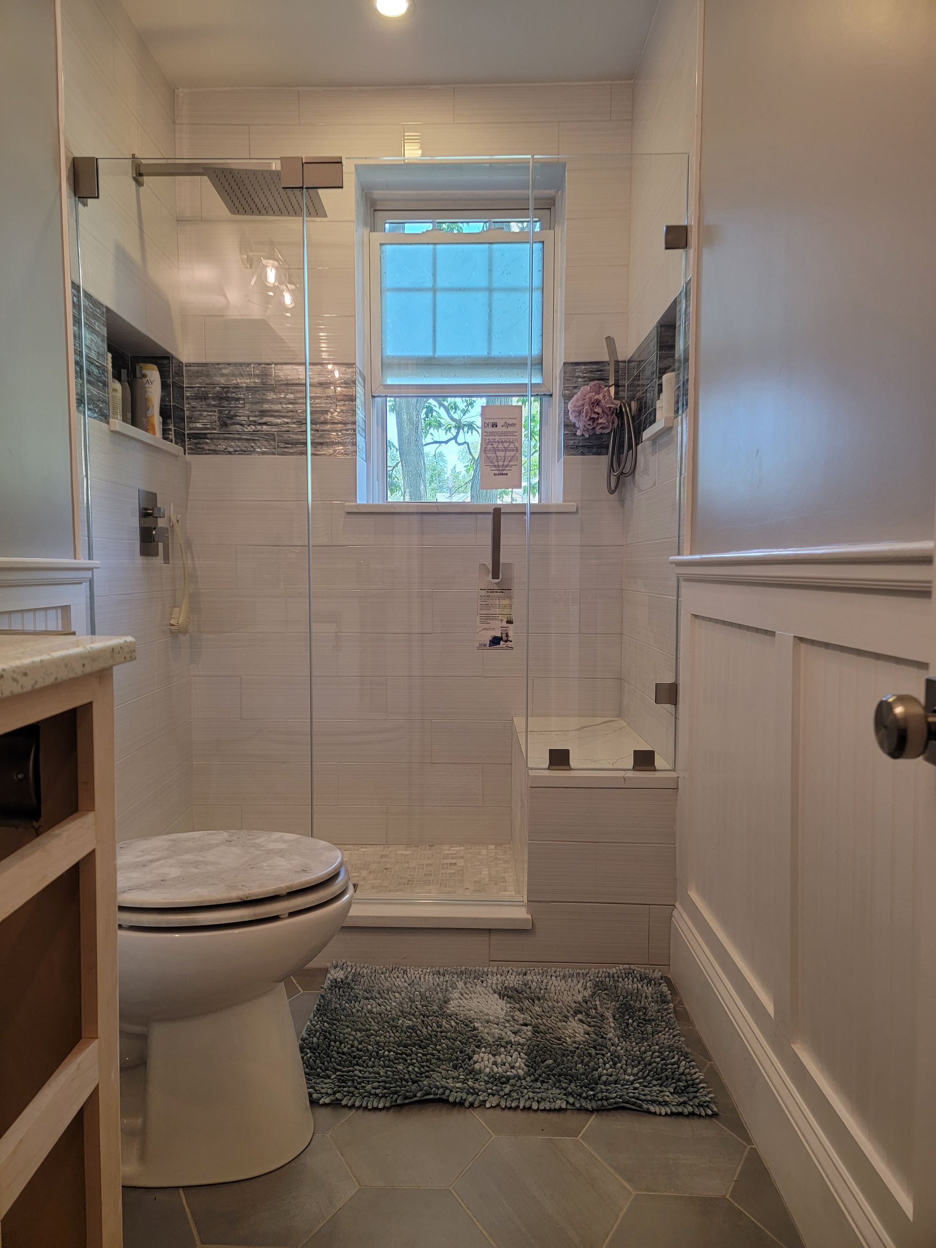 Small bathroom with white tiled shower, glass door, and toilet. Blue rug, window, and wood paneling visible.
