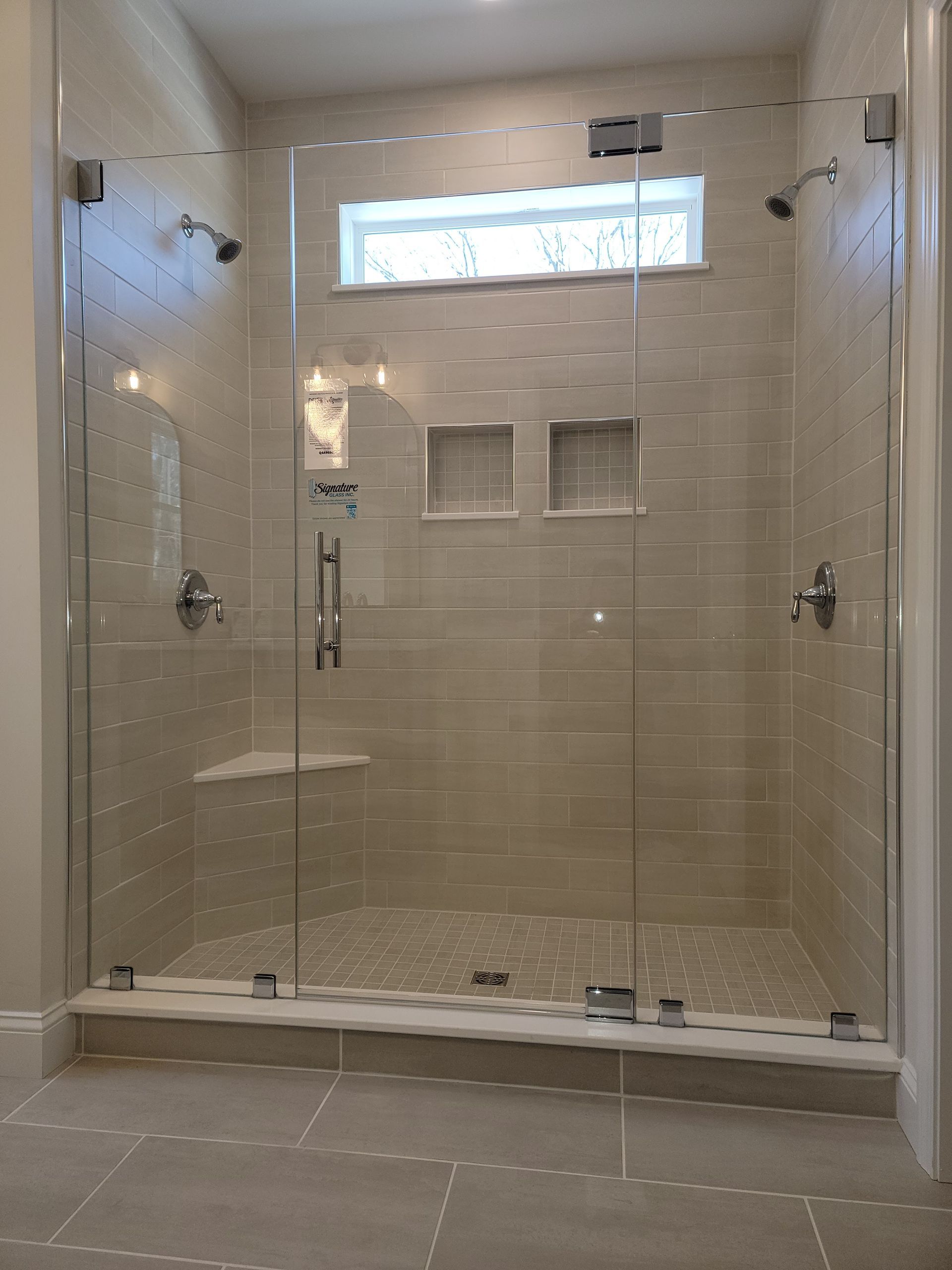 Glass-enclosed shower with tiled walls and pebble floor; two showerheads, a seat, and a window.