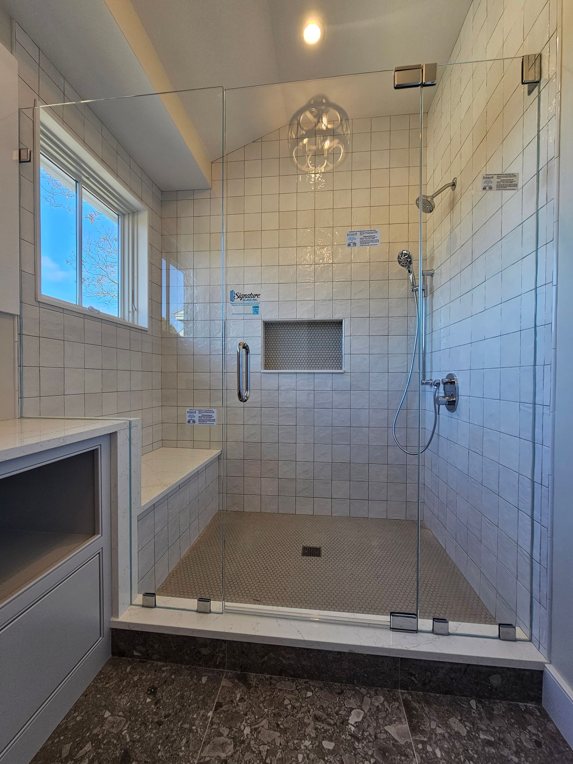 Glass-enclosed shower with stone floor and mosaic tile walls, built-in bench, and a window.