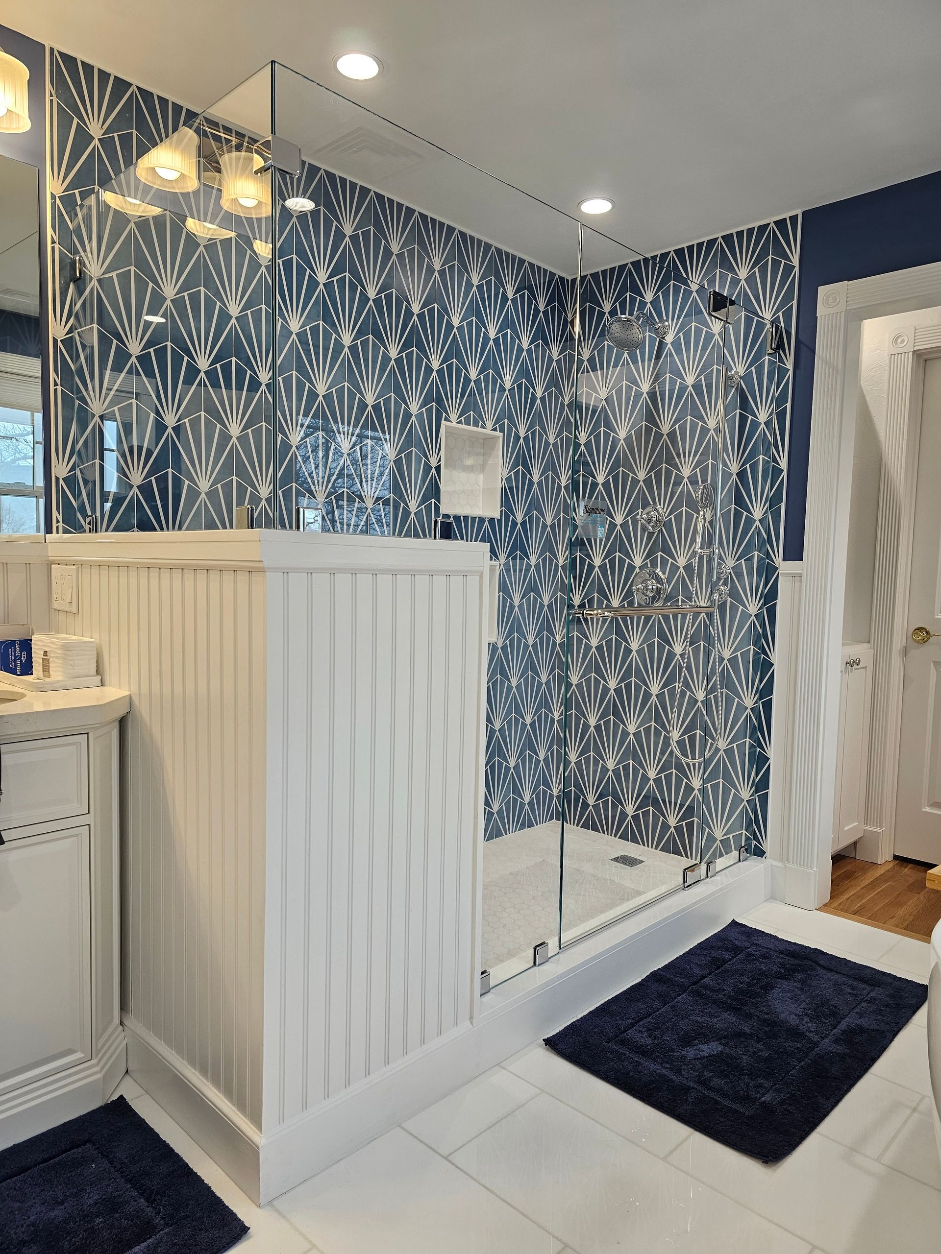 Bathroom with patterned blue tile shower, white wainscoting, glass door, and blue rug.