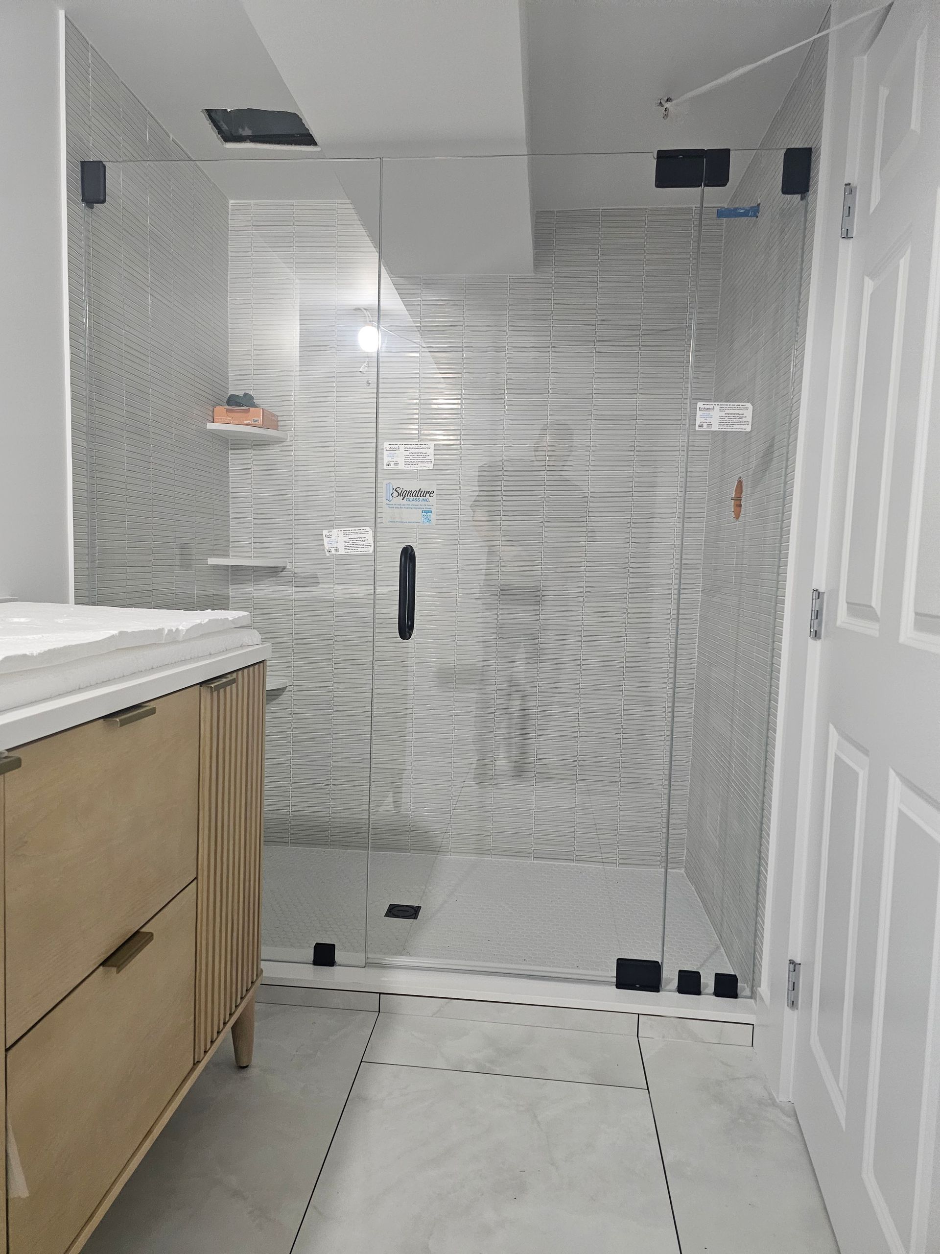 Bathroom with glass shower enclosure and light-colored vanity. Gray and white tile wall. Door on right.