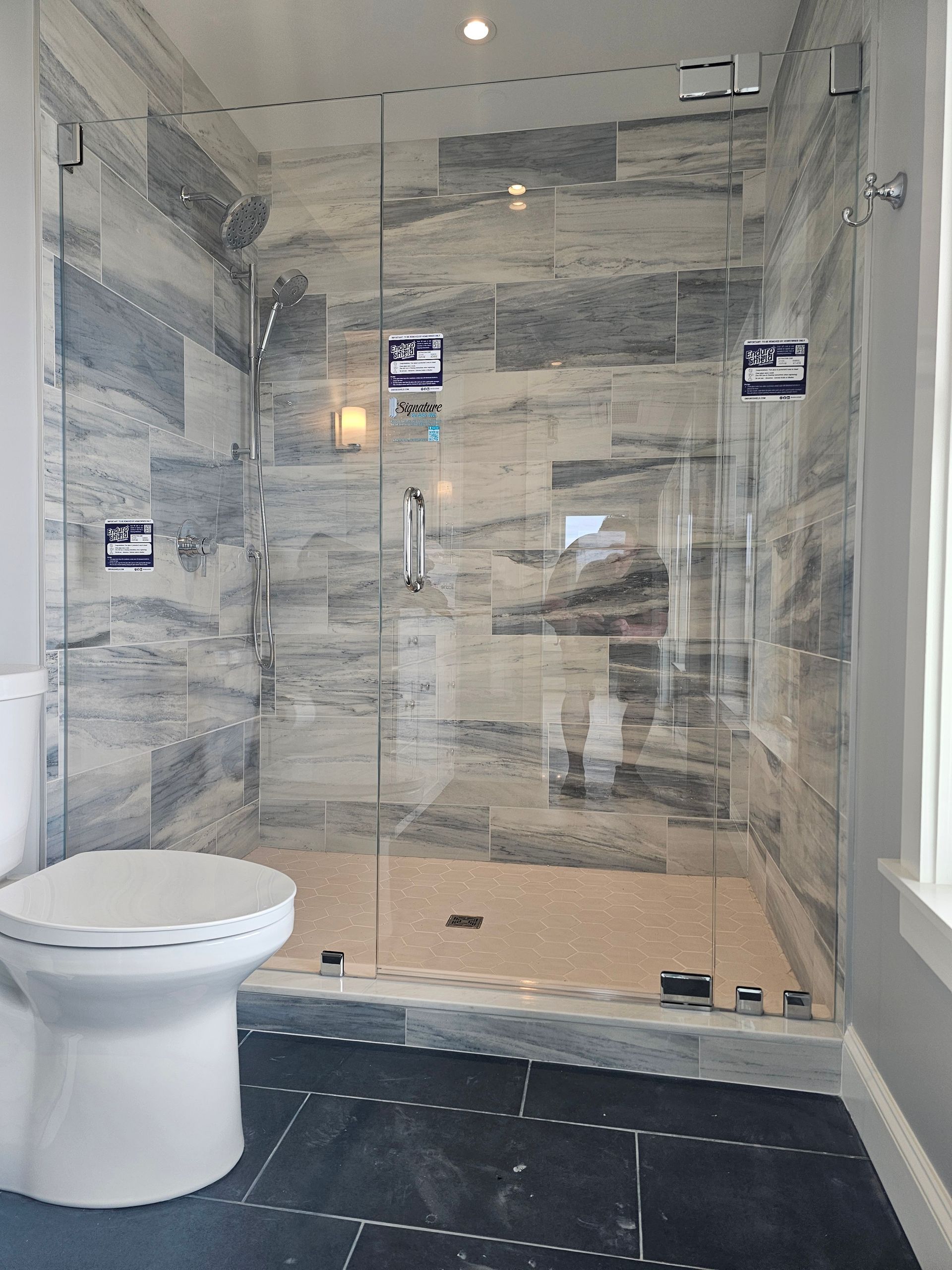 Shower with glass doors and grey tile walls, next to a white toilet on black floor tiles.