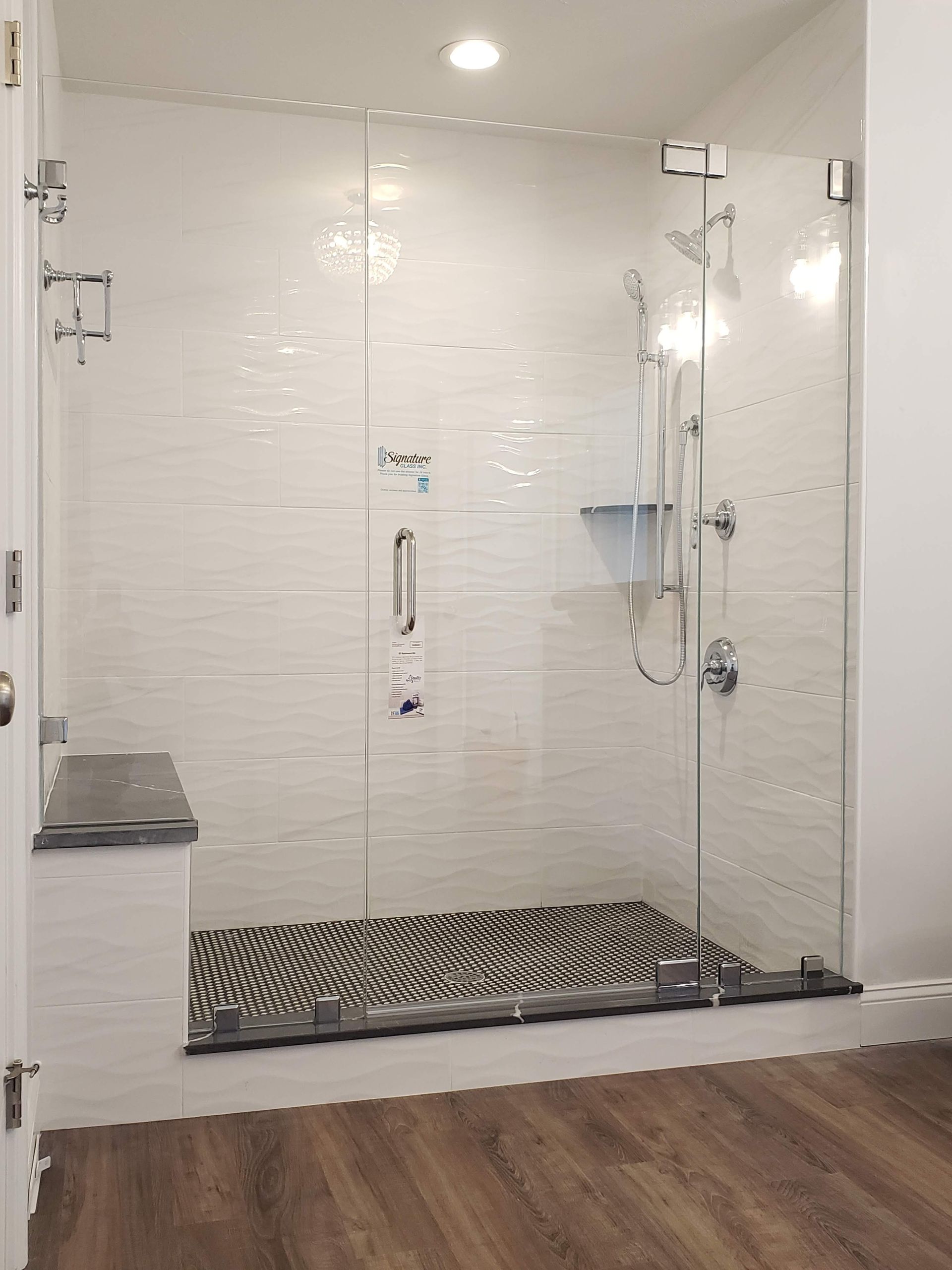 A modern glass shower with white tiled walls, pebble floor, and wooden flooring outside.
