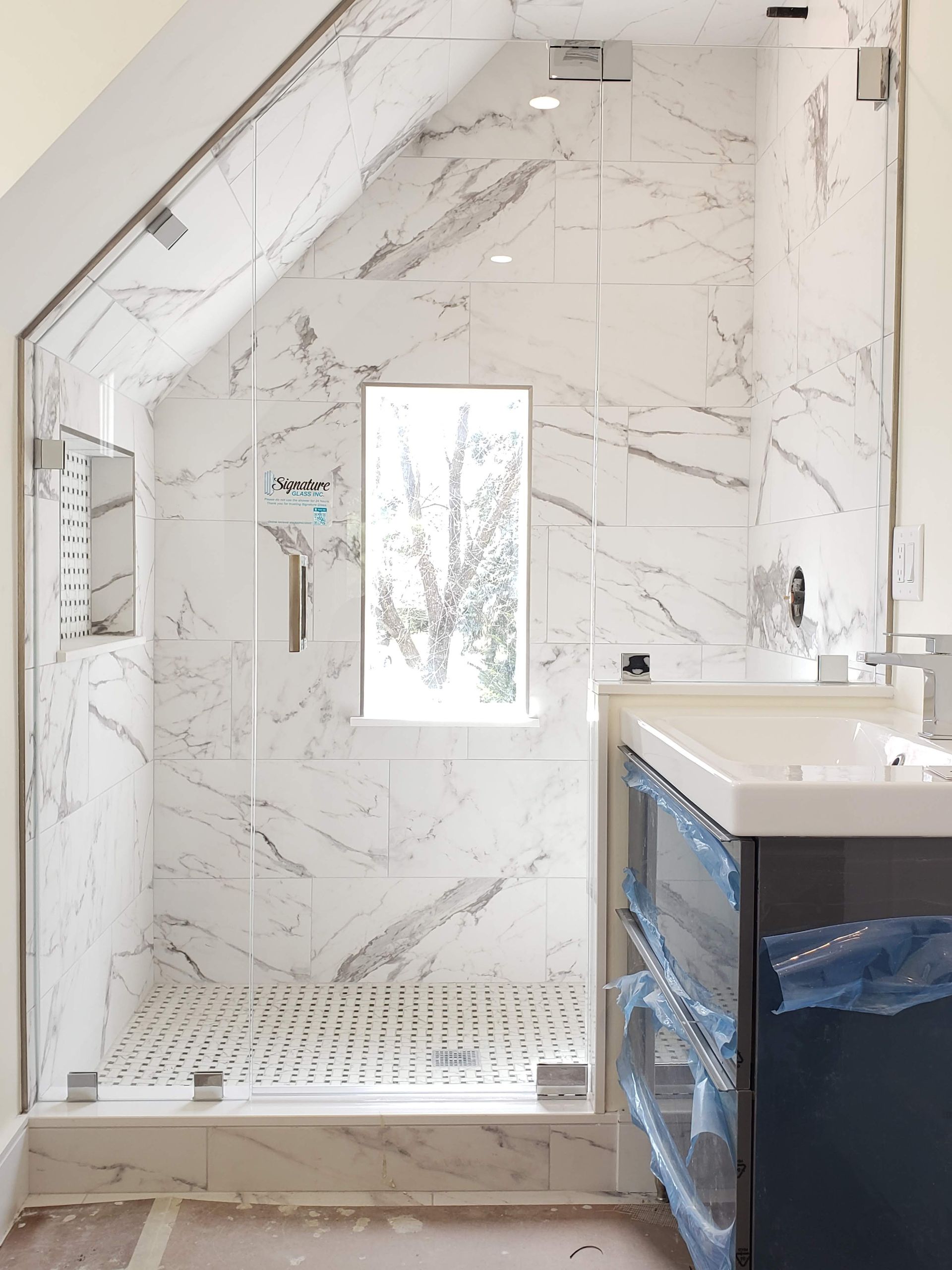 Shower with marble-patterned tiles, glass door, and white floor. A window is in the shower's back wall. A blue cabinet with a white countertop is on the right.
