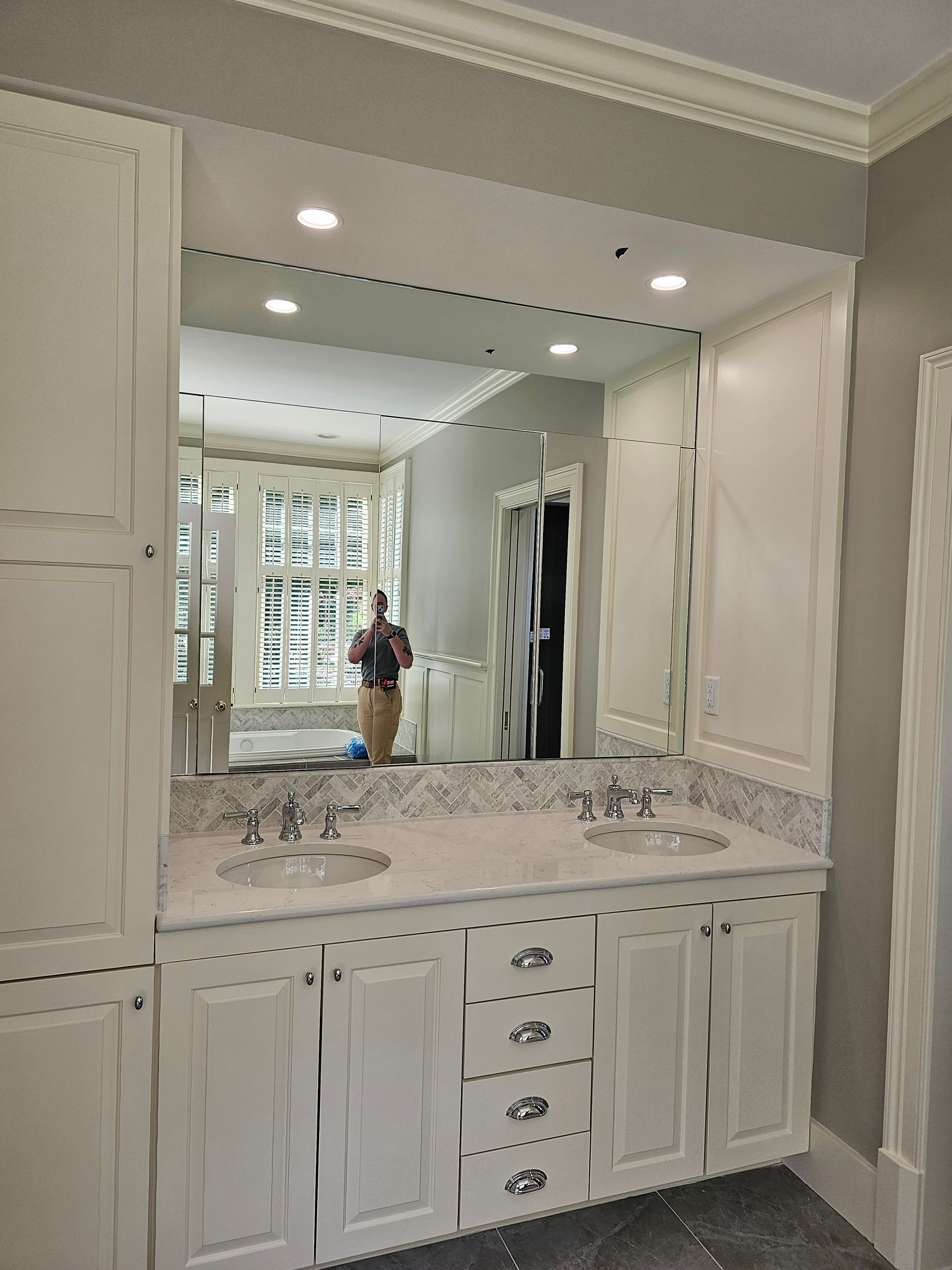 Bathroom vanity with white cabinets, marble countertop, large mirror, and person reflected.