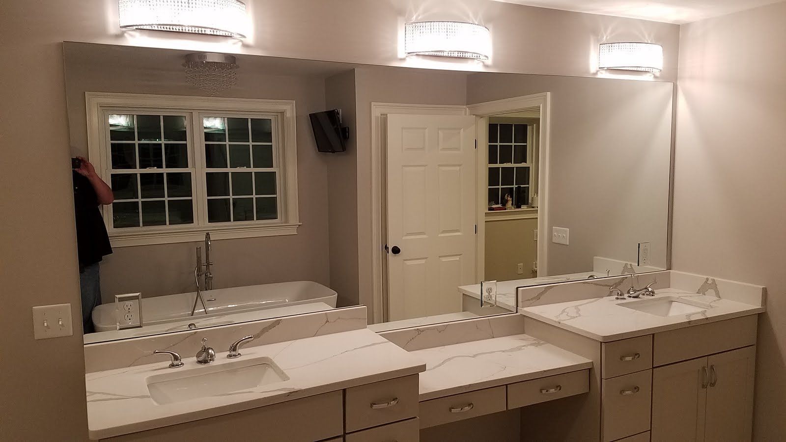 Bathroom with large mirror, vanity, sinks, lights, window with tub view, person adjusting mirror.