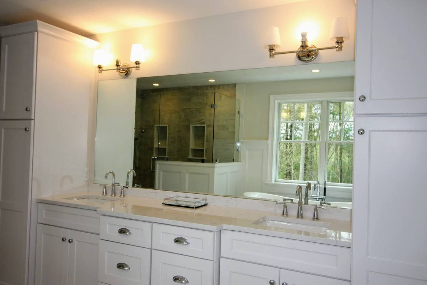 Bathroom with white cabinets, large mirror, double sink, and a window overlooking a wooded area.