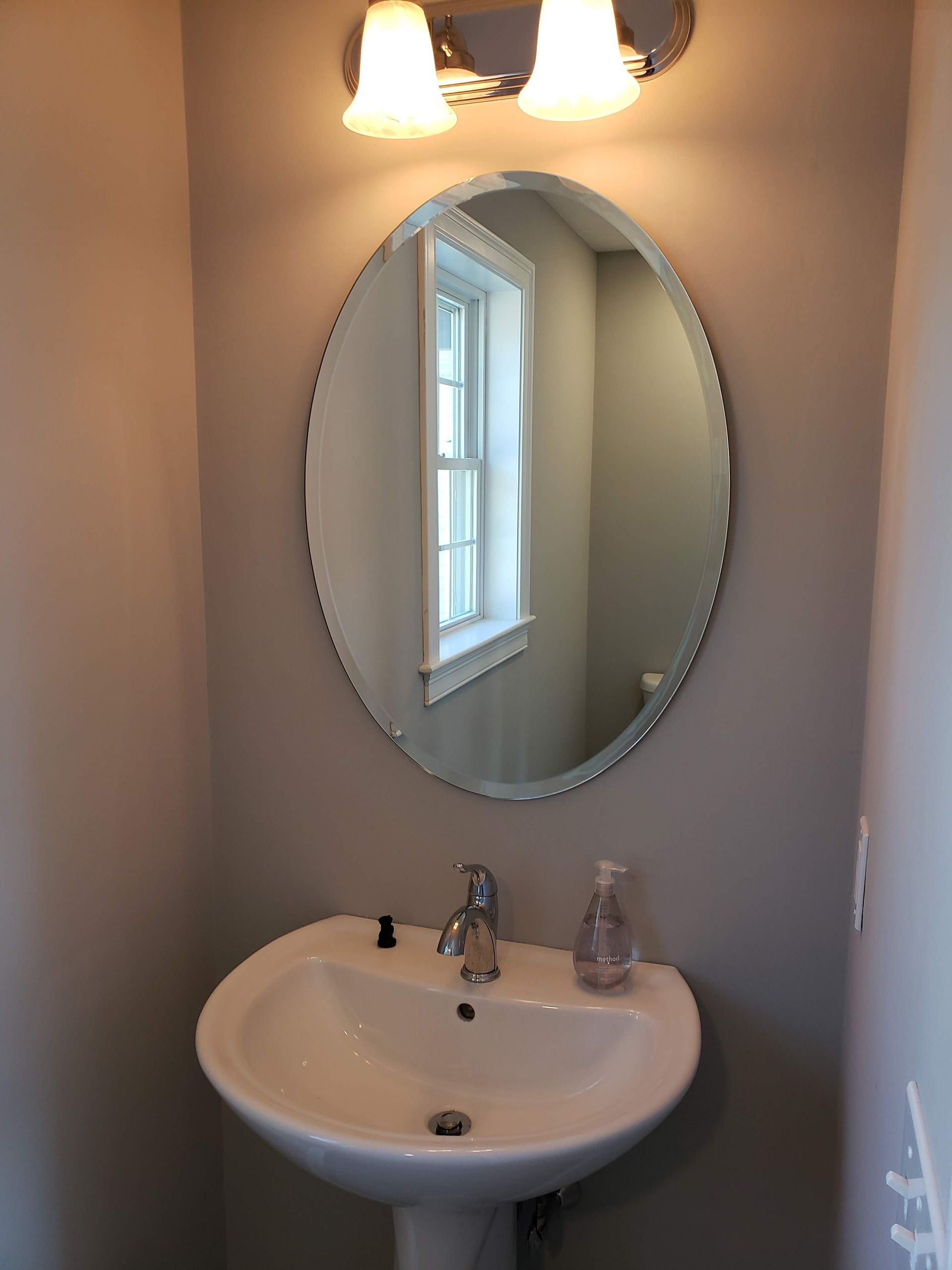 Bathroom with a pedestal sink, oval mirror, and a window reflecting the outside. Gray walls.