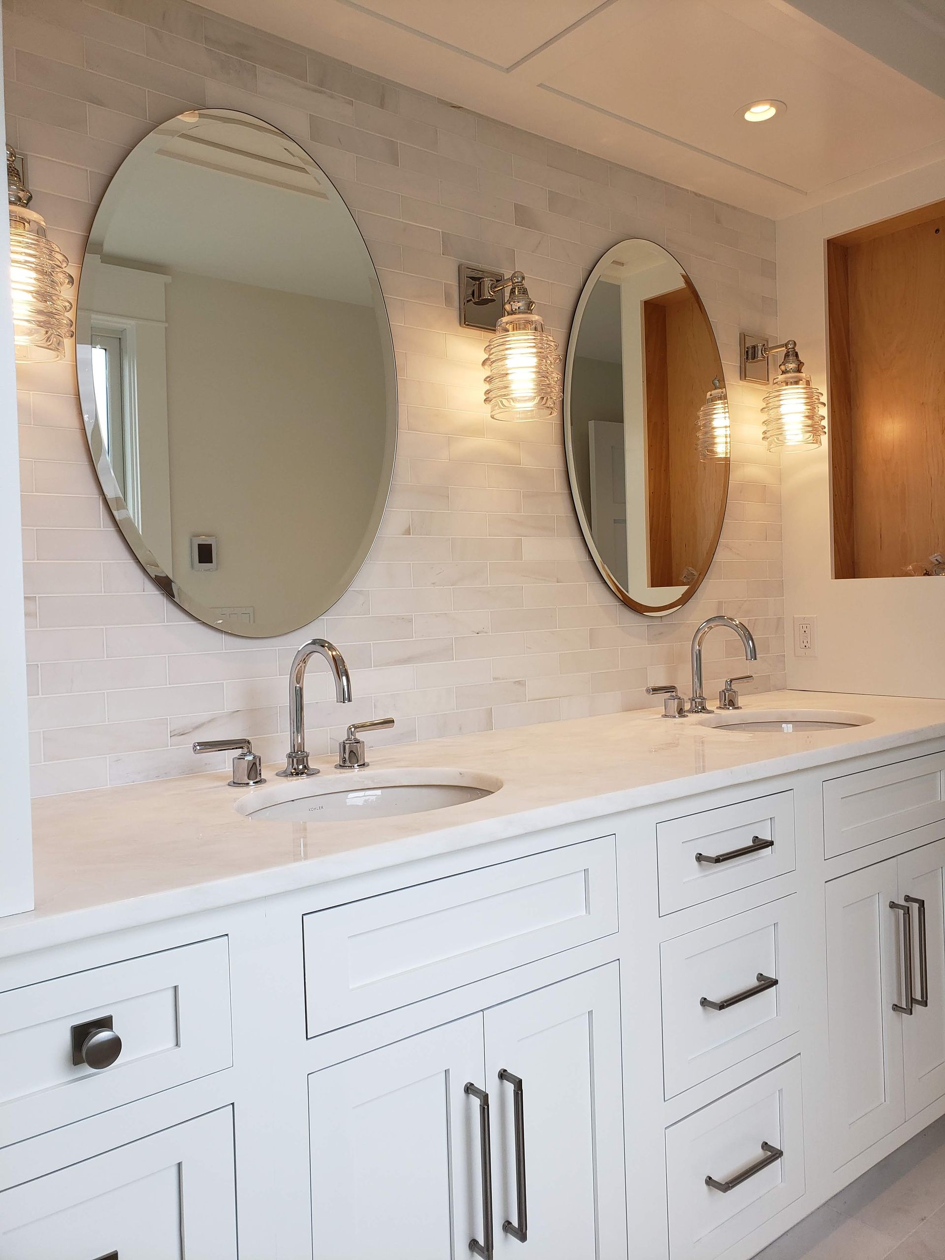 White bathroom vanity with oval mirrors, sconces, and brushed silver faucets.