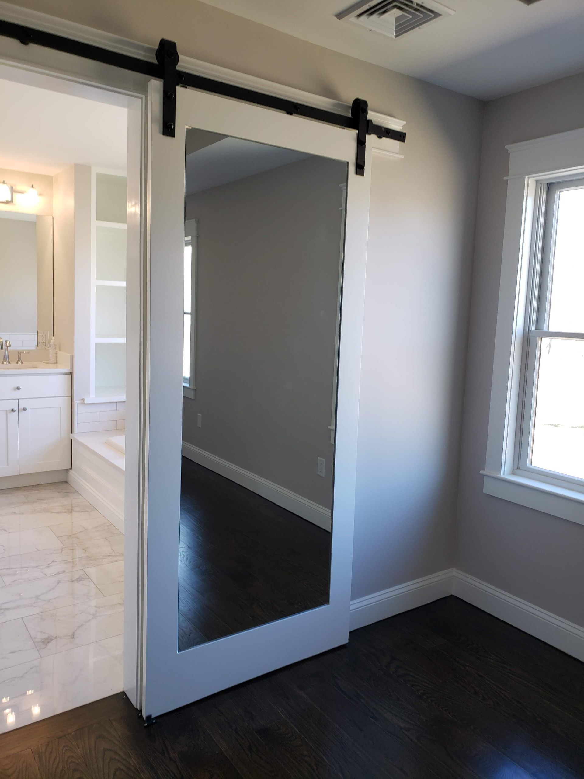 White sliding barn door with full-length mirror, black hardware, dark wood floor, light gray walls, open to bathroom.