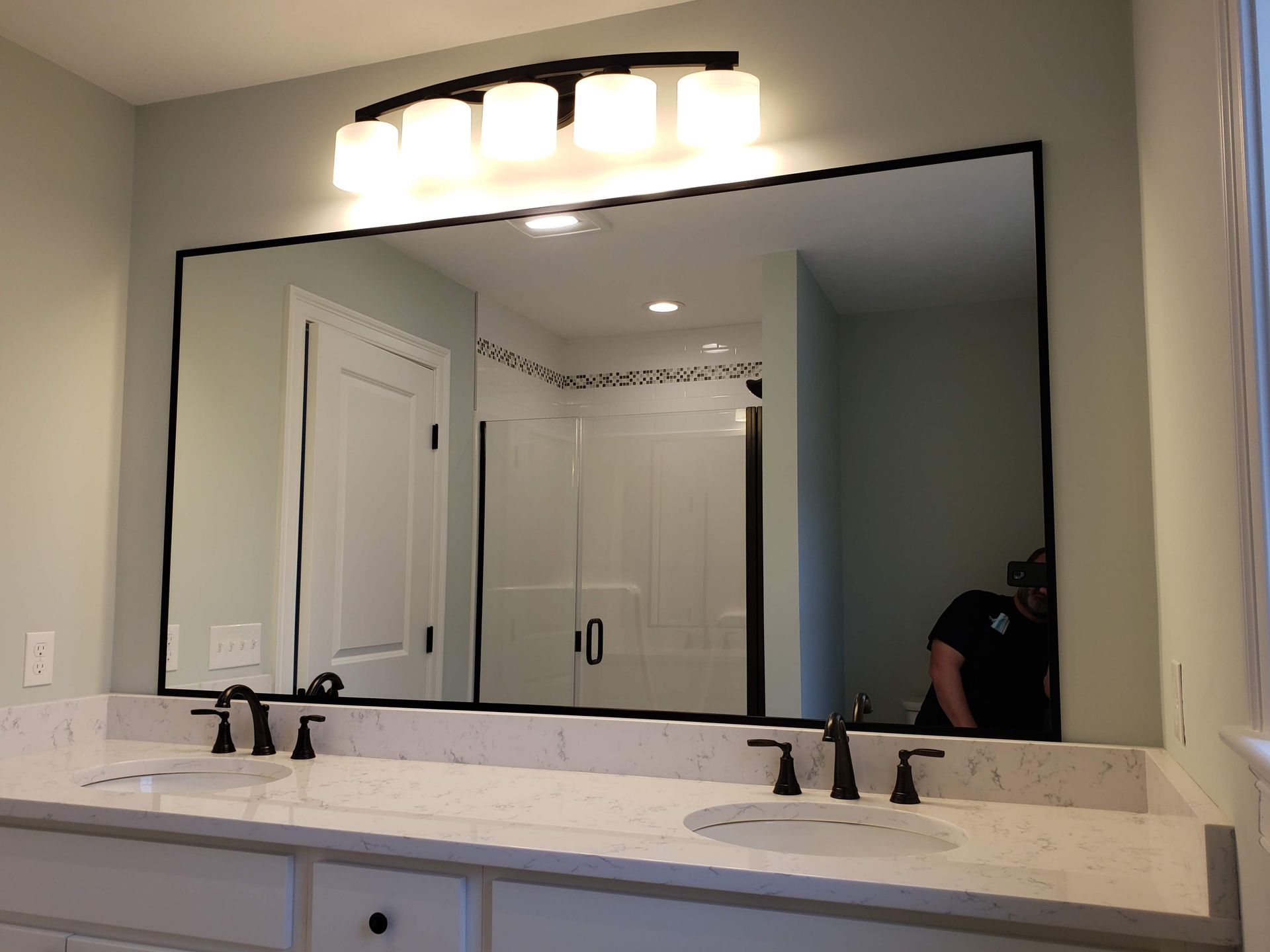 Bathroom with a large mirror above a double sink vanity. Black framed mirror, light fixture, and fixtures.