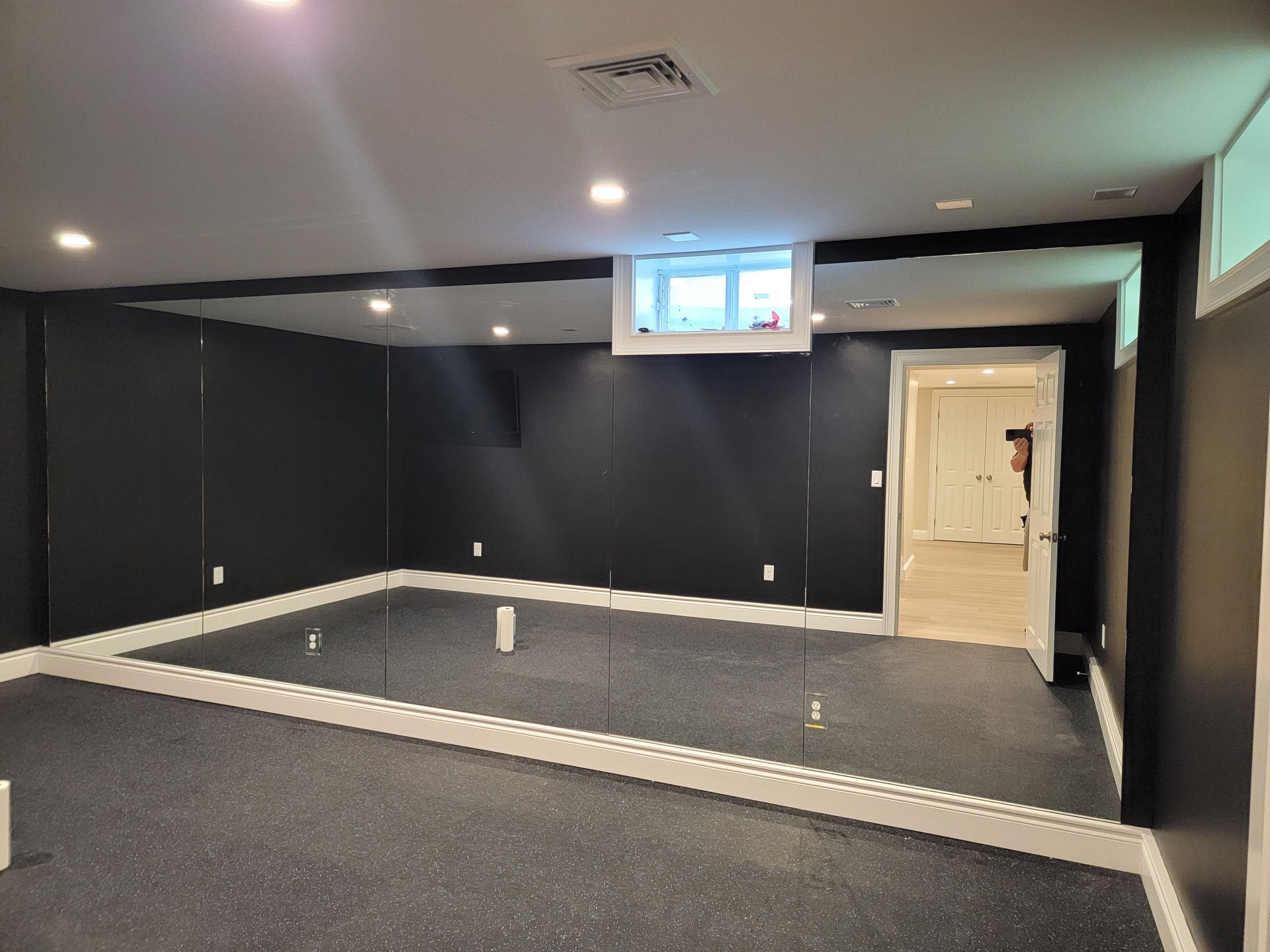 Black-walled room with a large mirror covering most of one wall, dark flooring, and a door to another room.