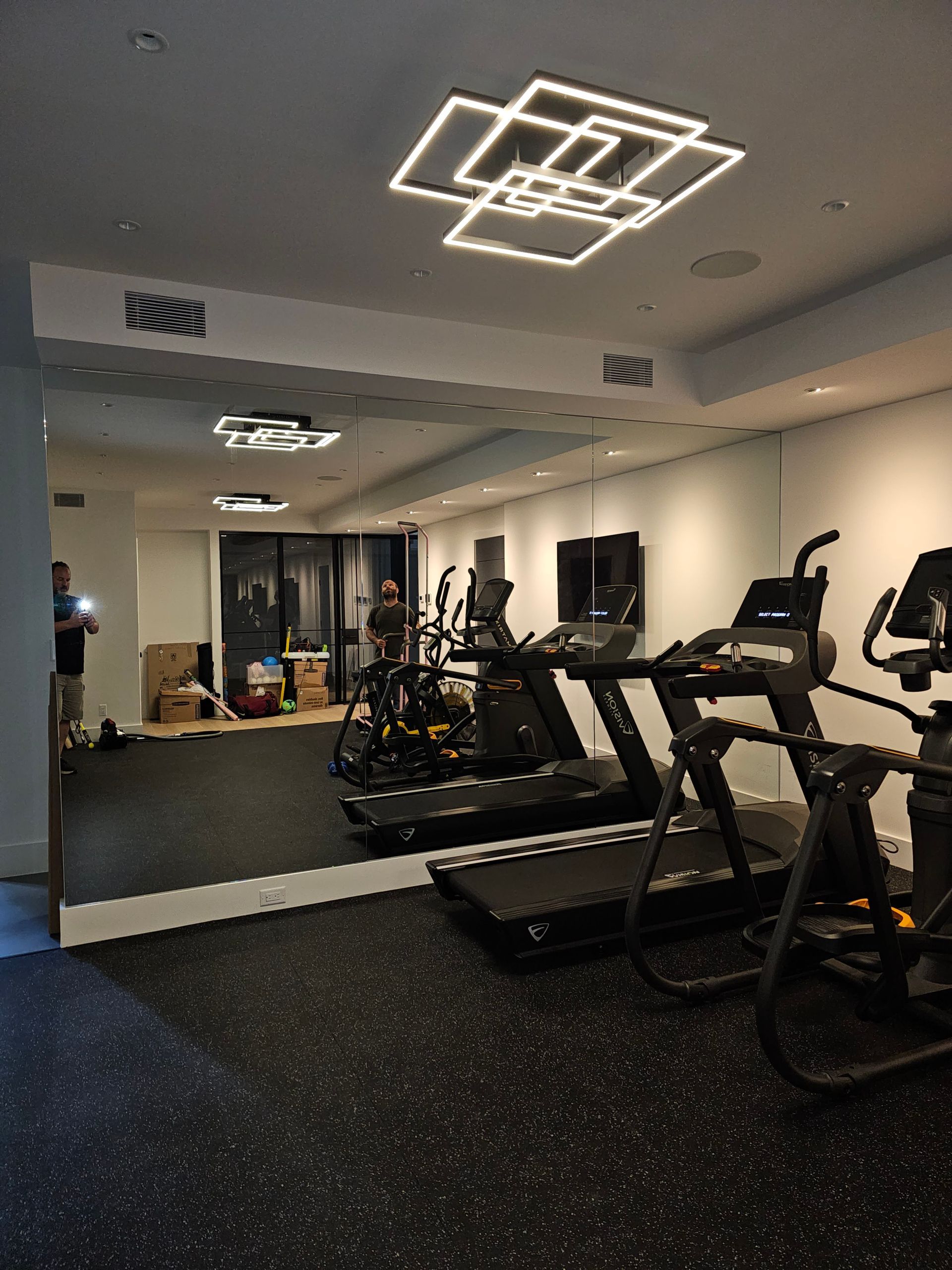 Gym with fitness equipment, including treadmills and elliptical machines, mirrored wall, modern lighting.