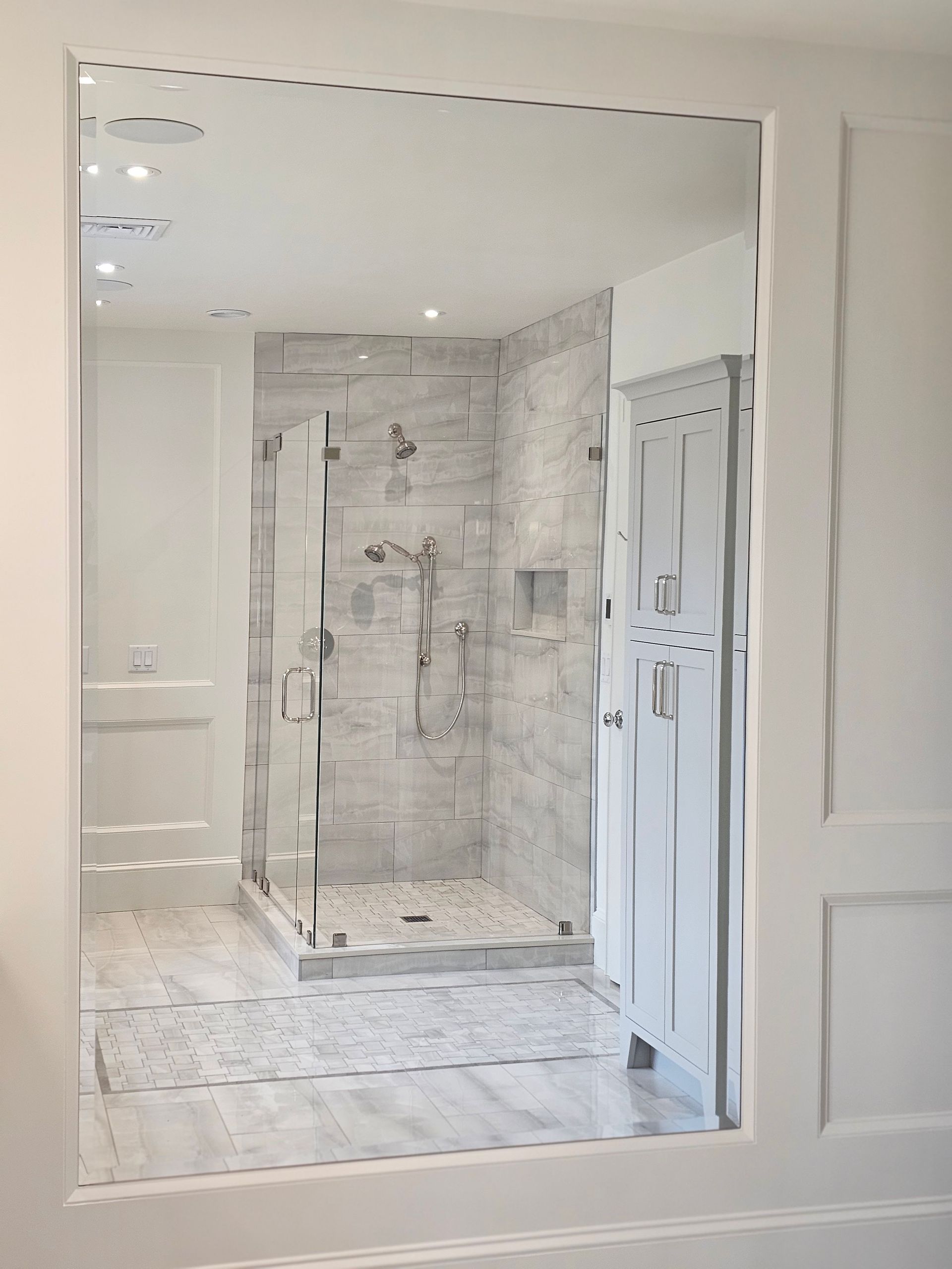 Bathroom interior with shower, tiled walls, and white cabinetry reflected in mirror.