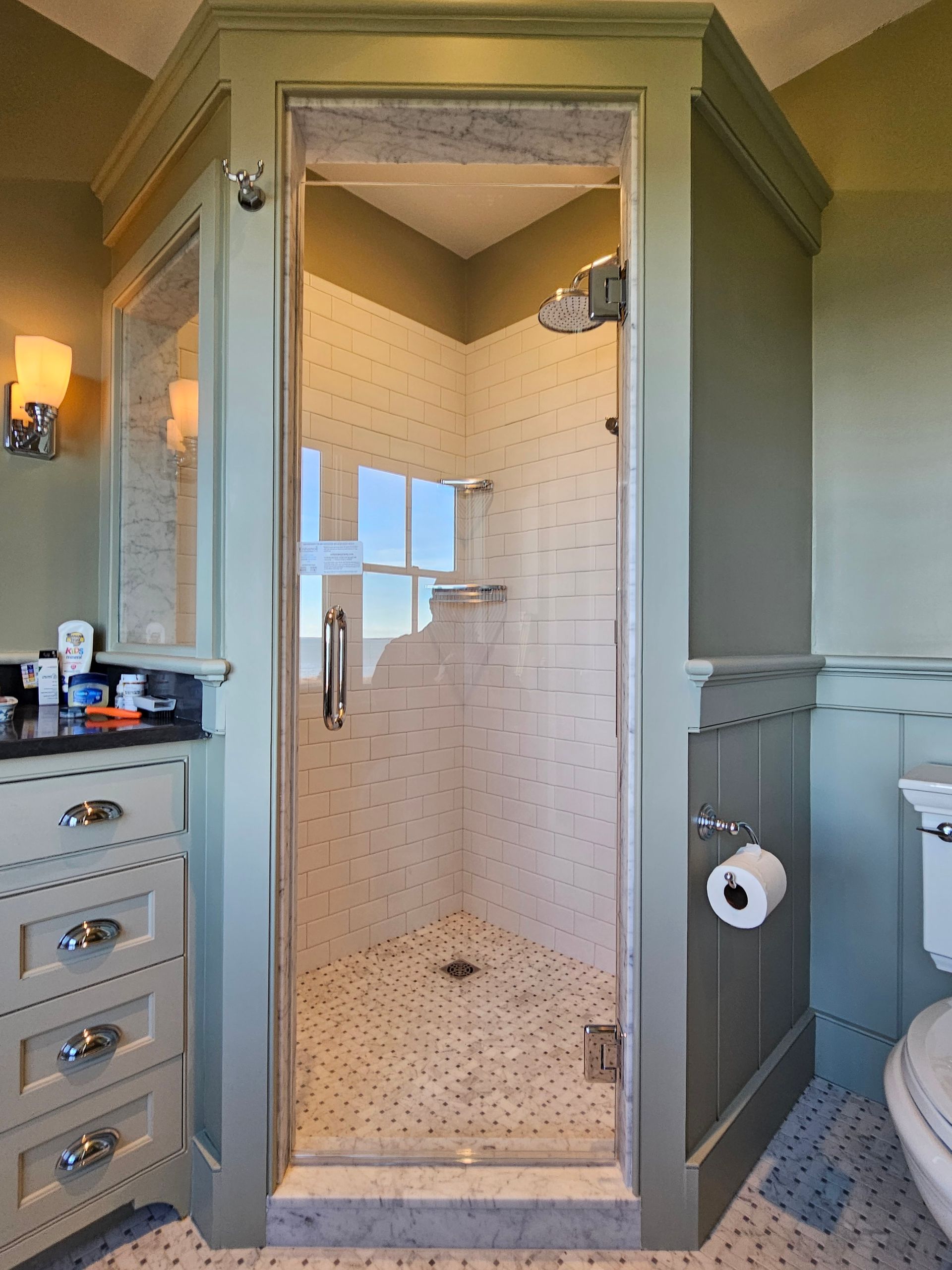 A walk-in shower with glass doors, white tiled walls, and a small window, set in a green-painted bathroom.