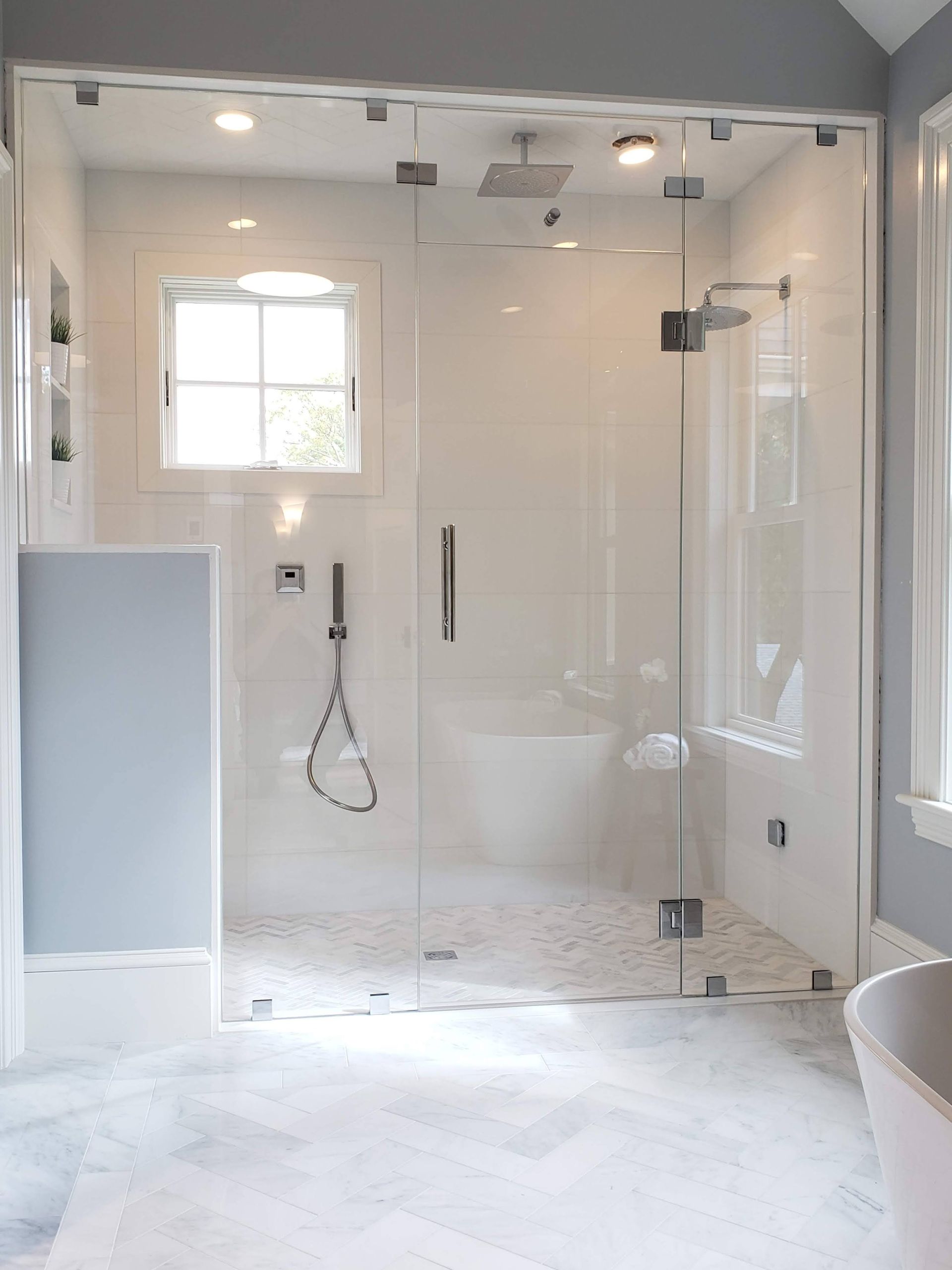 Glass-enclosed shower in a modern bathroom. White tile, rain showerhead, window, and marble flooring.