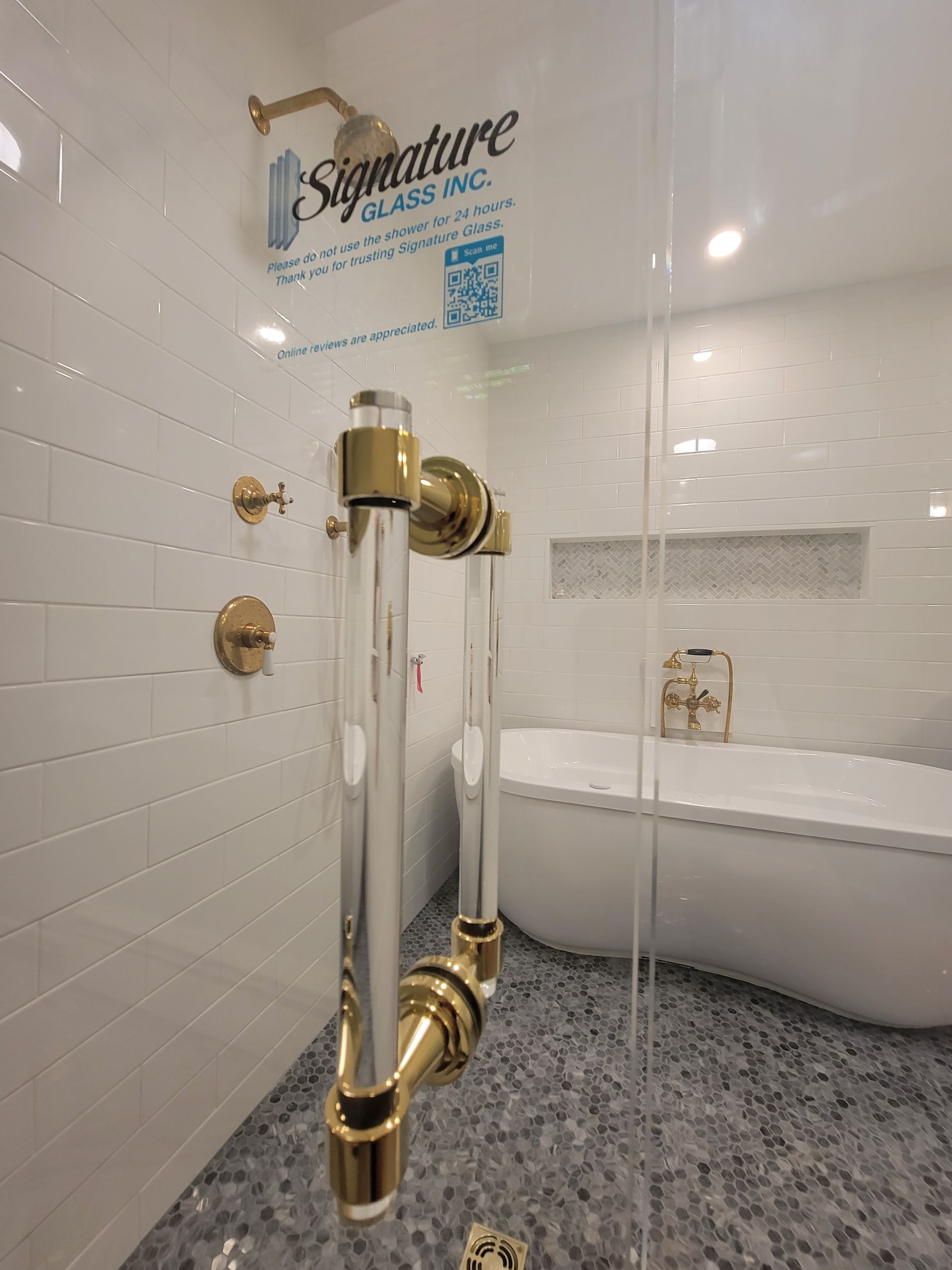 Gold and clear glass shower door with gold fixtures, white tiled walls, and a soaking tub.