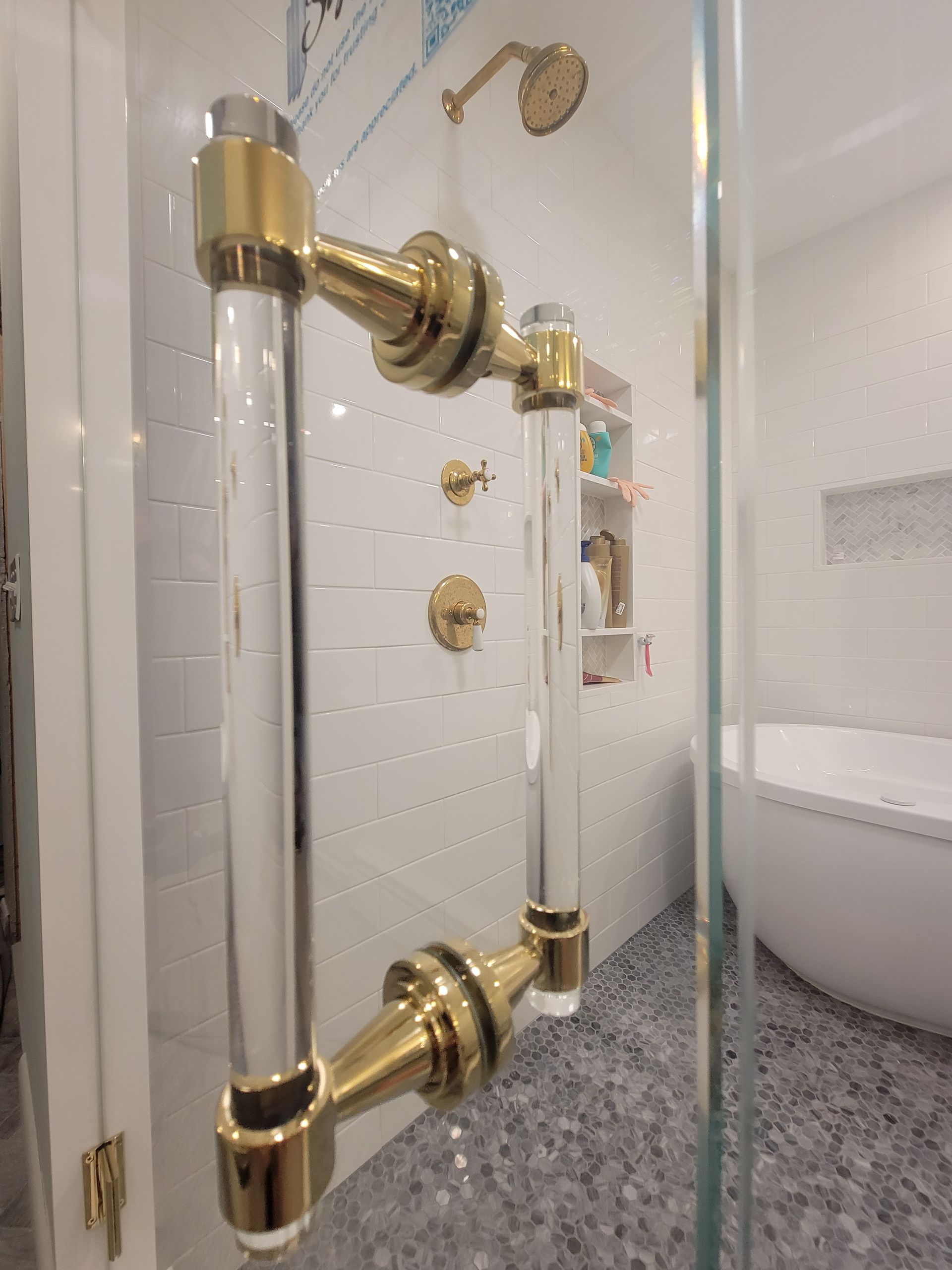 Clear shower door with gold and acrylic handles, gold shower fixtures, and white tiled wall.