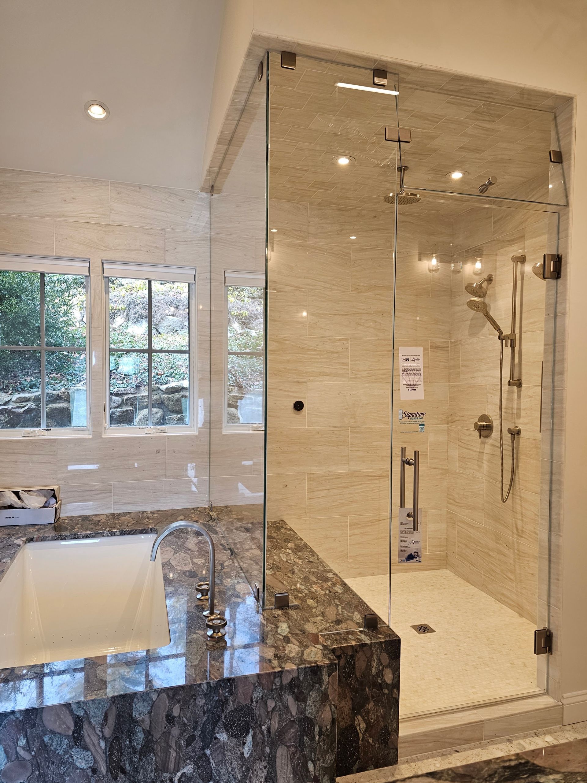 Modern bathroom with glass shower enclosure, marble countertop, windows, and light-colored stone walls.