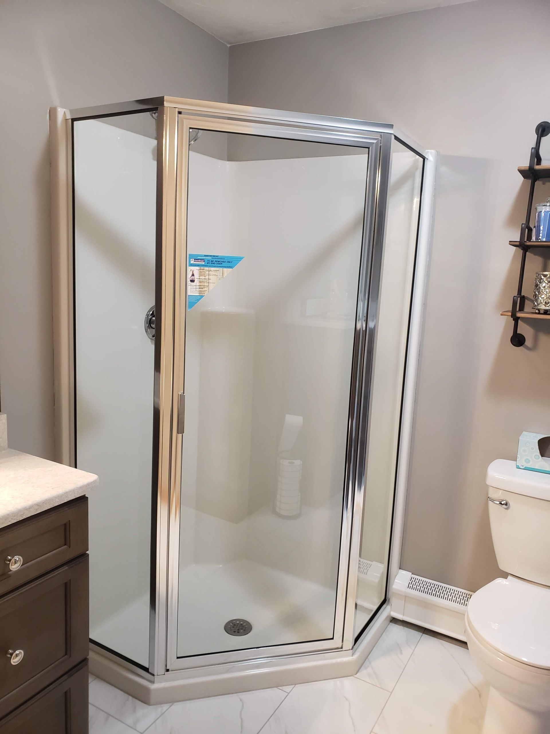 Corner shower with white walls, glass door, chrome trim, next to a toilet and vanity.